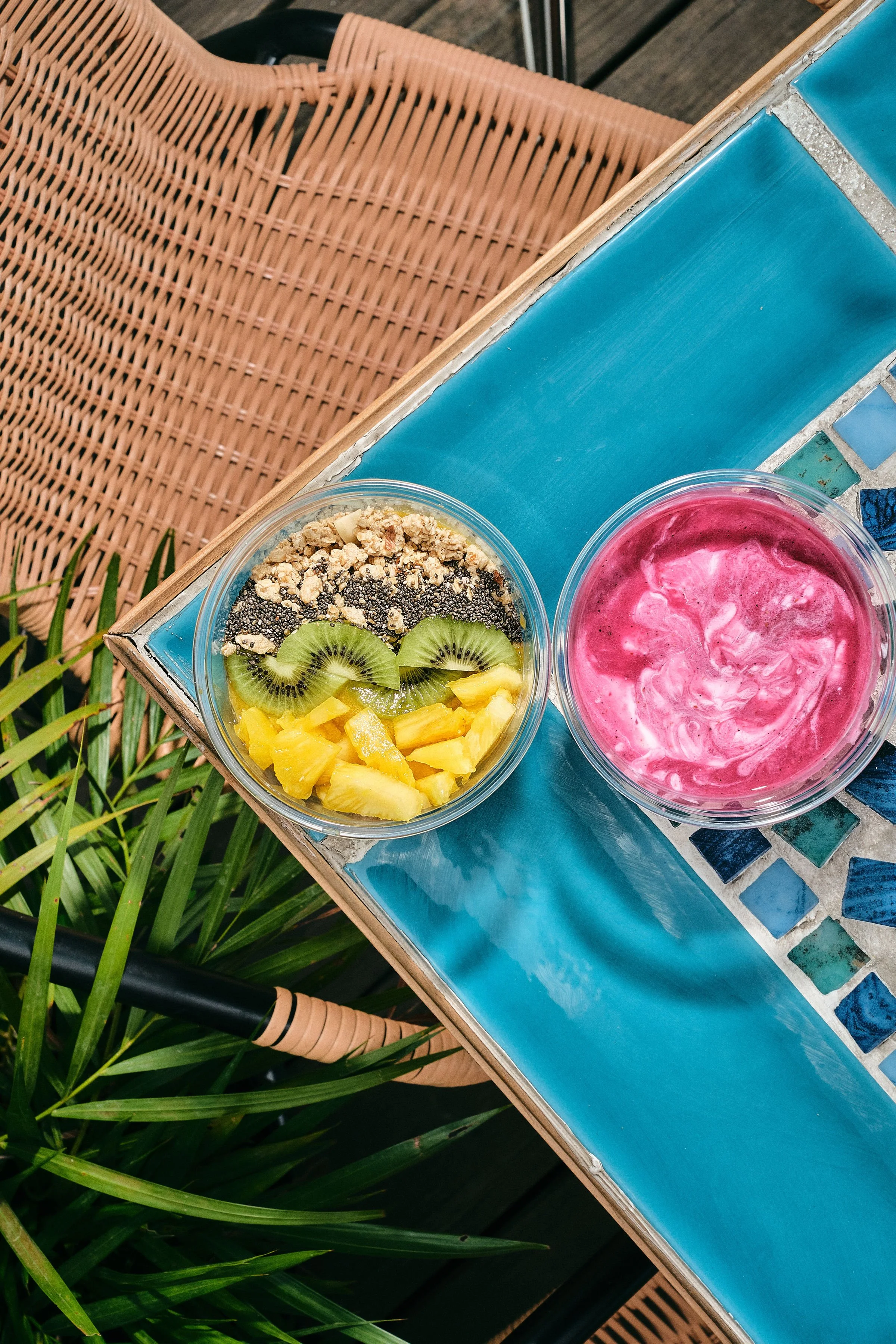 Two bowls of colorful smoothies and fruit toppings on a blue-tiled outdoor table, with a rattan chair and green plants nearby.