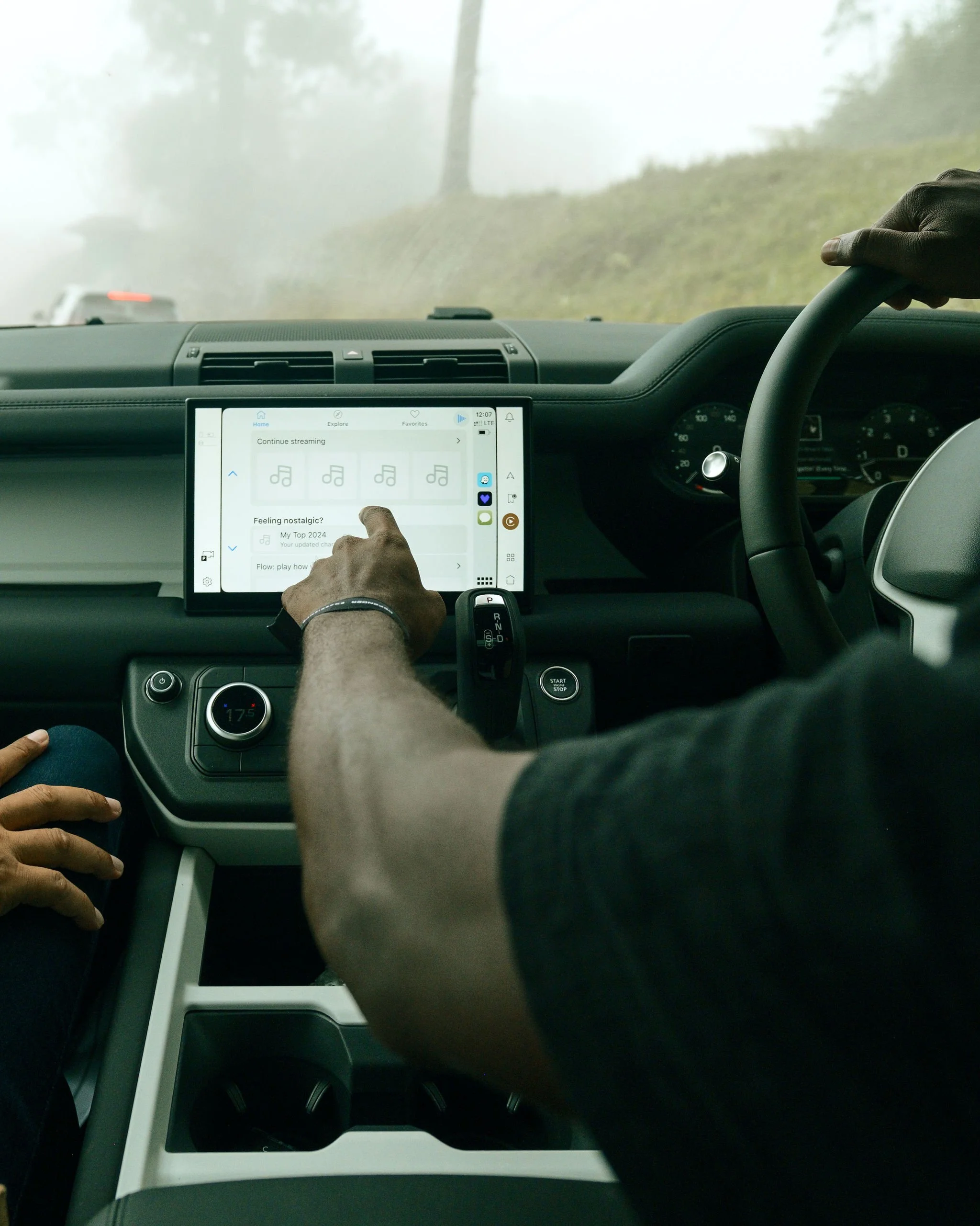 Inside a vehicle, a person's hand is touching a touchscreen displaying music options, with foggy outside scenery visible through the windshield.