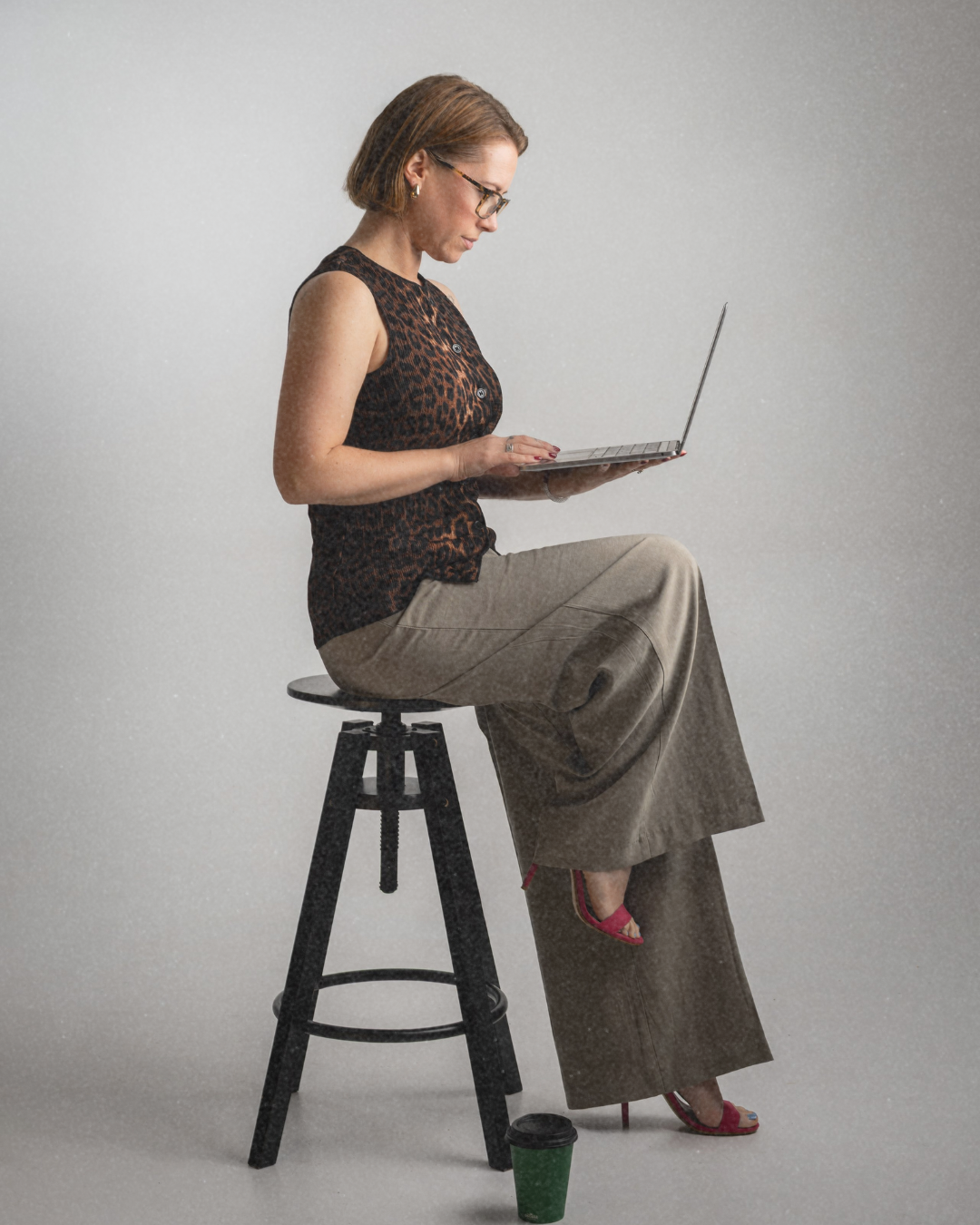 A woman sitting on a black stool, looking at a laptop in her hands, against a plain gray background, with a green takeaway coffee cup on the floor.