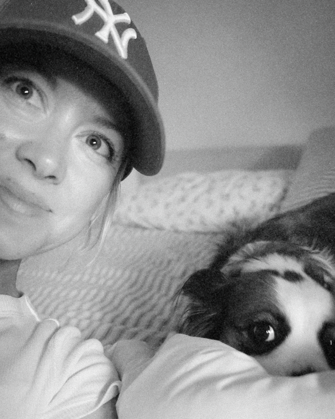 A woman in a baseball cap taking a selfie with her dog resting its head on a pillow, both lying on a bed.