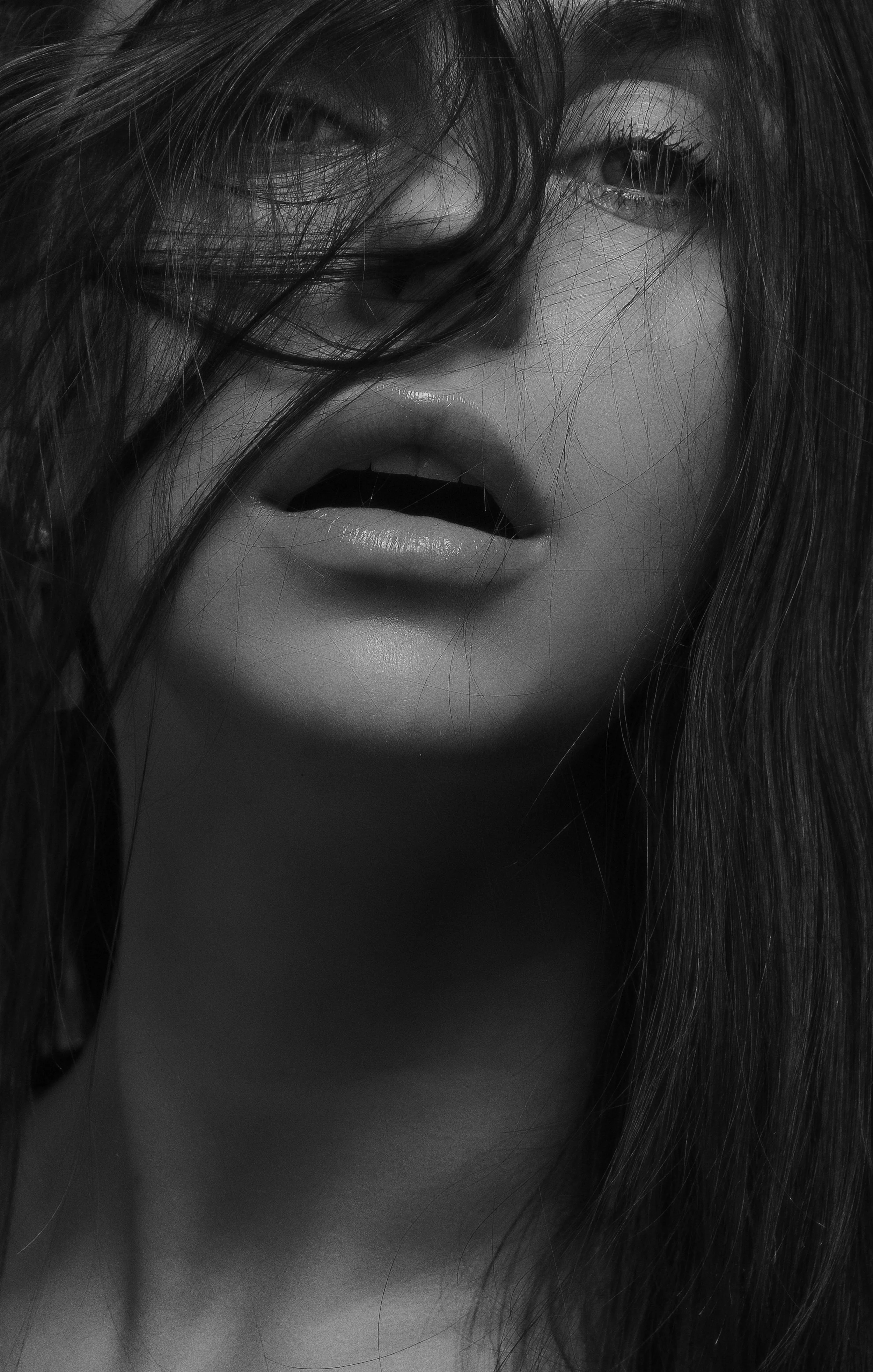 Close-up black and white photo of a woman with messy hair partially covering her face, mouth slightly open, and intense gaze.