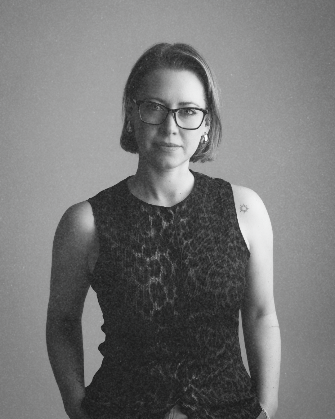 Black and white portrait of a woman with glasses, short hair, and earrings, wearing a sleeveless top, standing against a plain background.