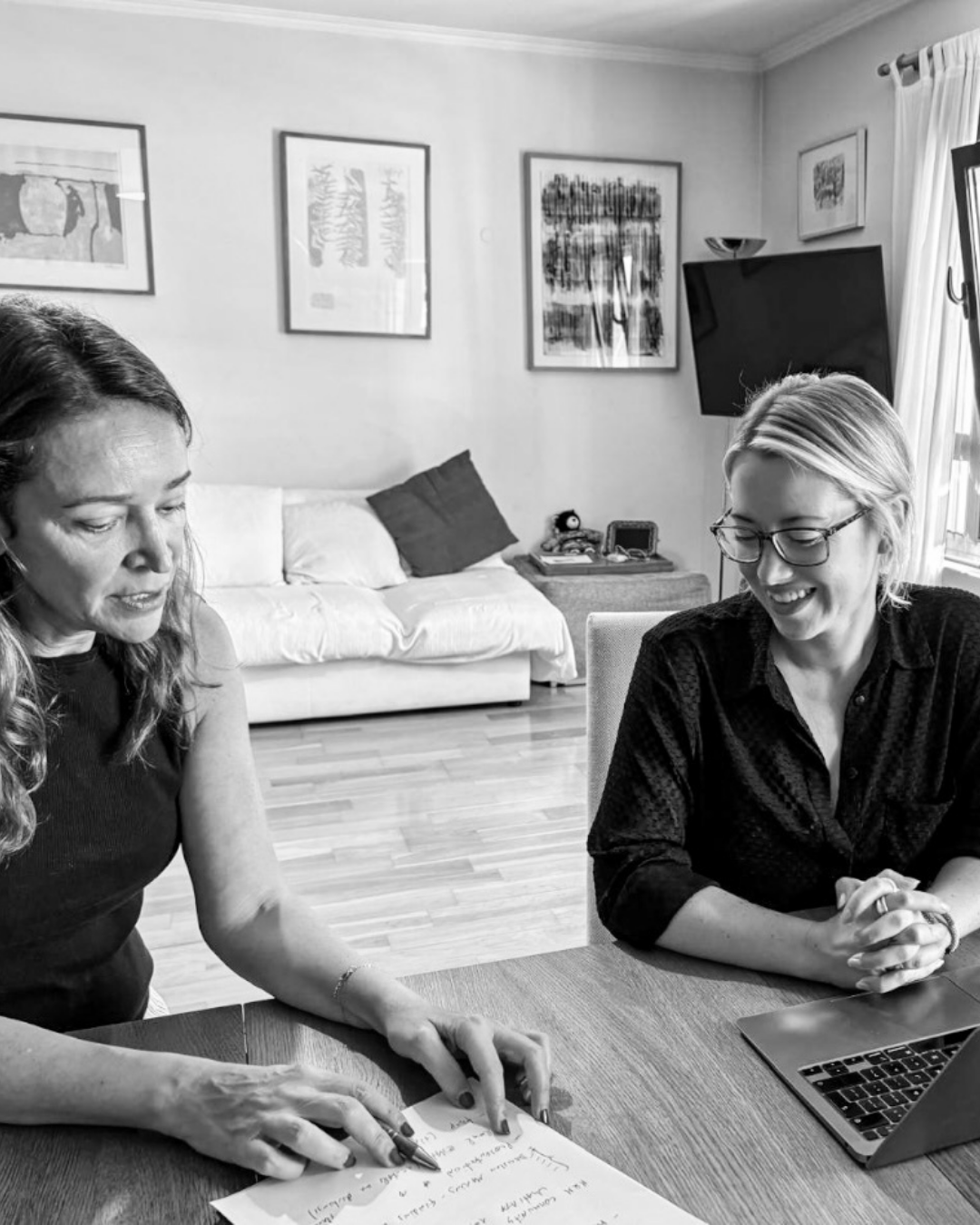 Two women sitting at a table, one pointing at handwritten notes and the other smiling, in a living room with artwork on the wall, a sofa, and a TV.