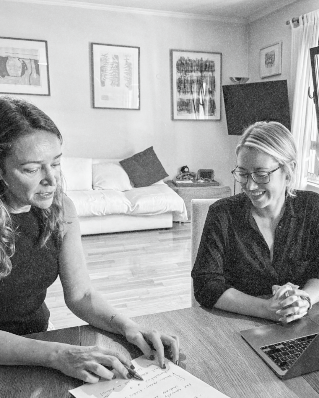 Two women sitting at a table with a laptop and notes, smiling and engaging in a discussion in a living room.