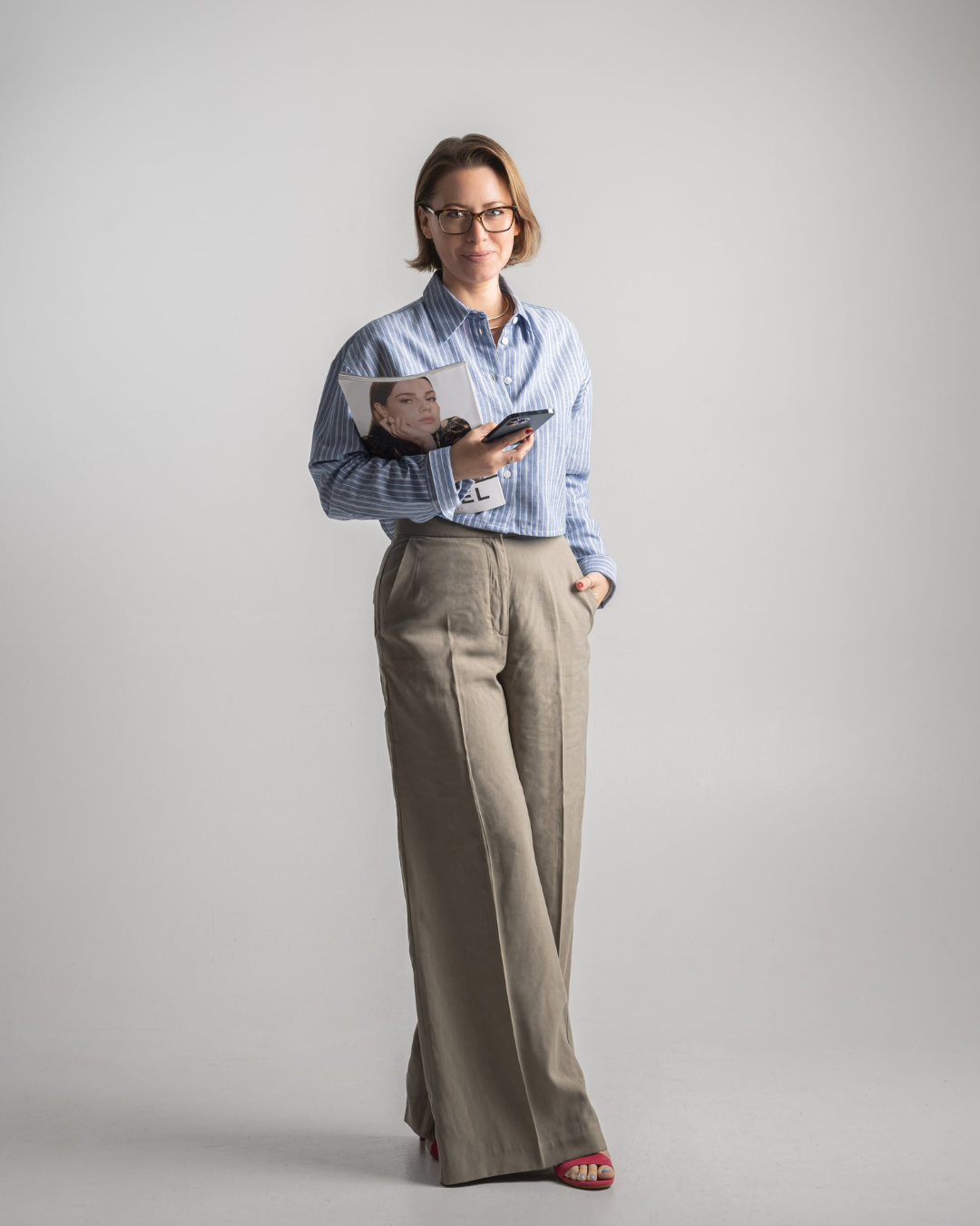 A woman in glasses, wearing a blue striped shirt and beige high-waisted trousers, holding a magazine and smartphone, standing against a plain white background.