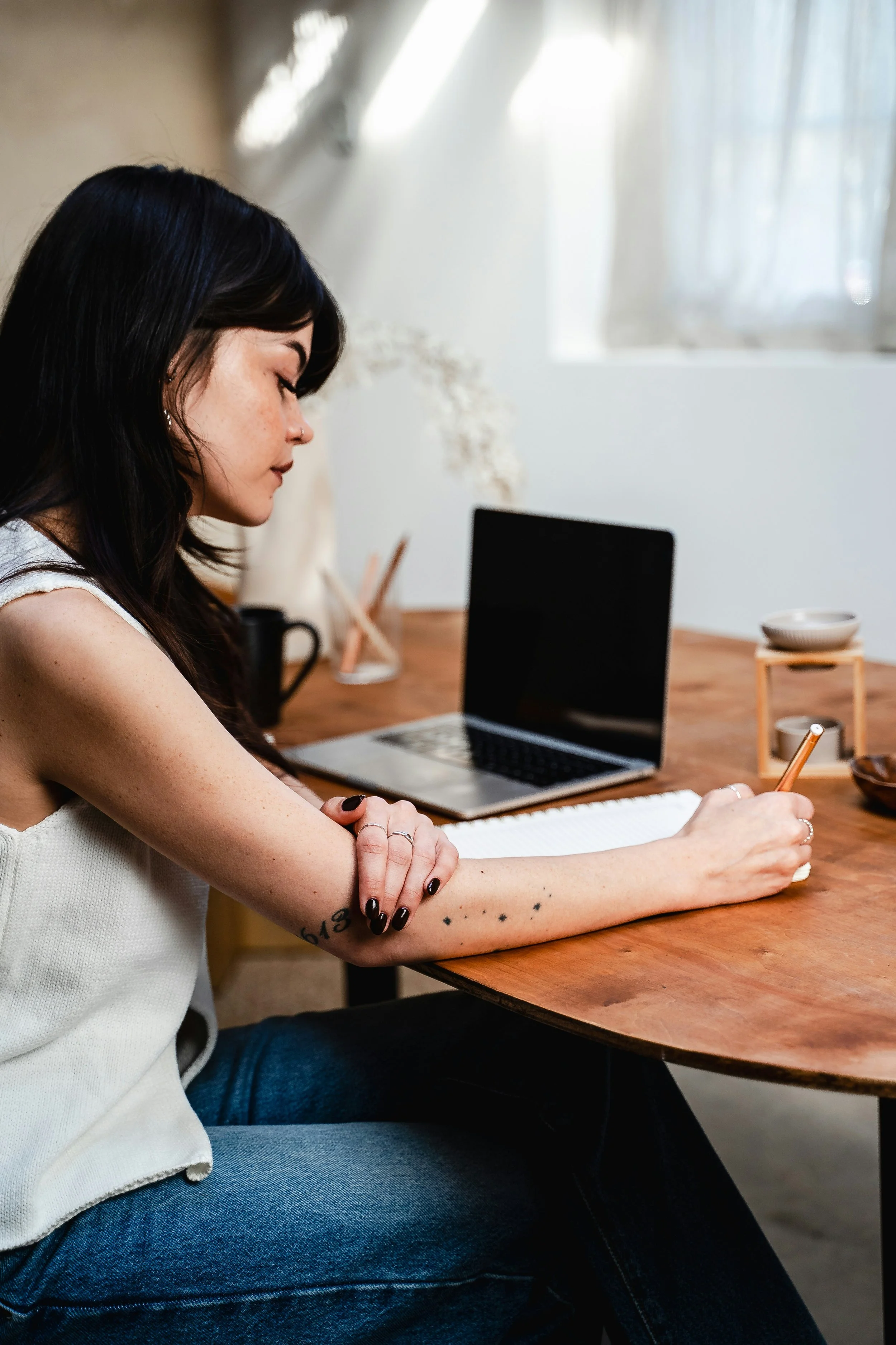 A woman with black hair and tattoos, sitting at a wooden table, writing in a notebook with a laptop open in front of her, surrounded by minimalist decor and natural light from a window.