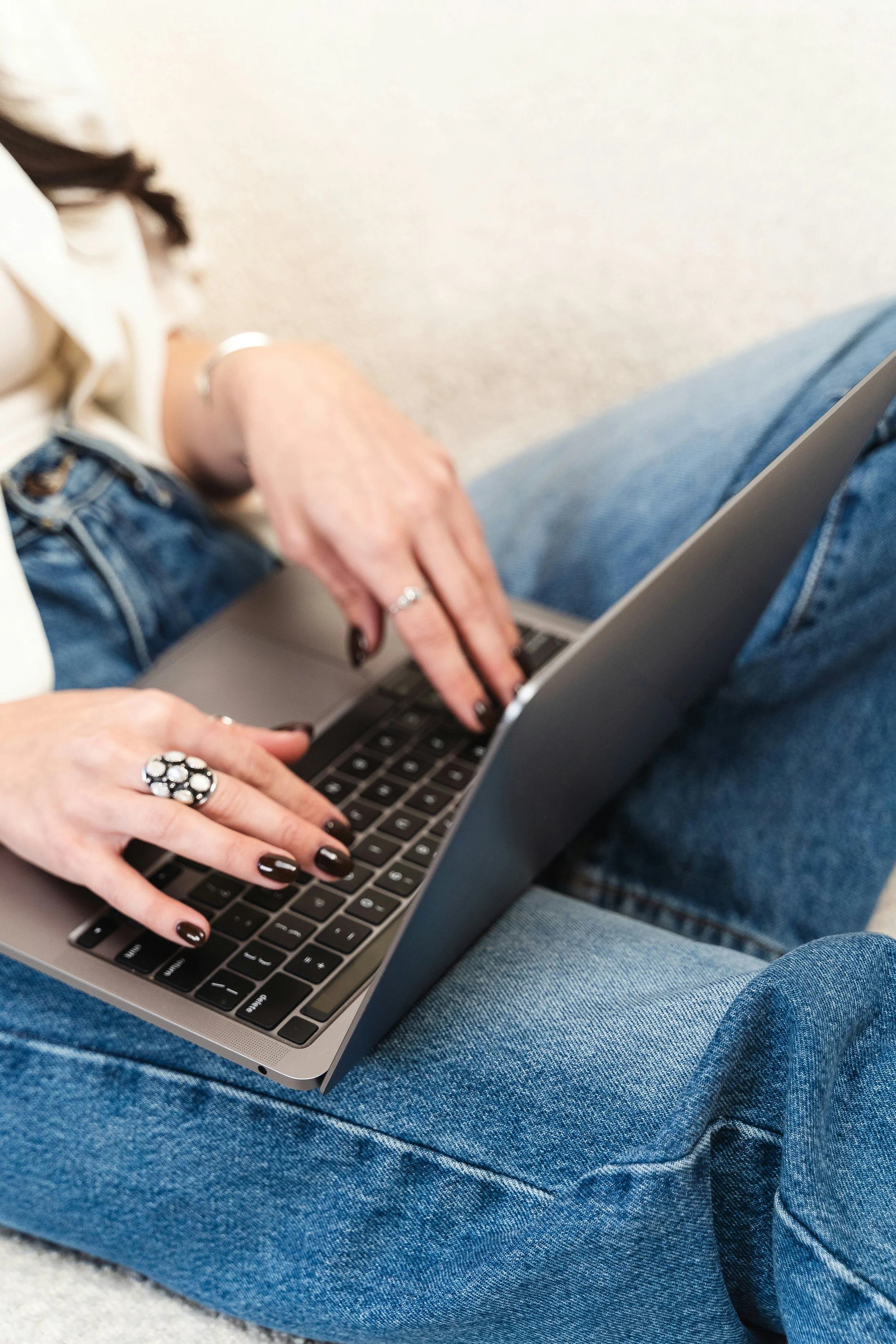 A person with black painted nails, wearing jewelry, typing on a laptop while sitting cross-legged on the floor in blue jeans.