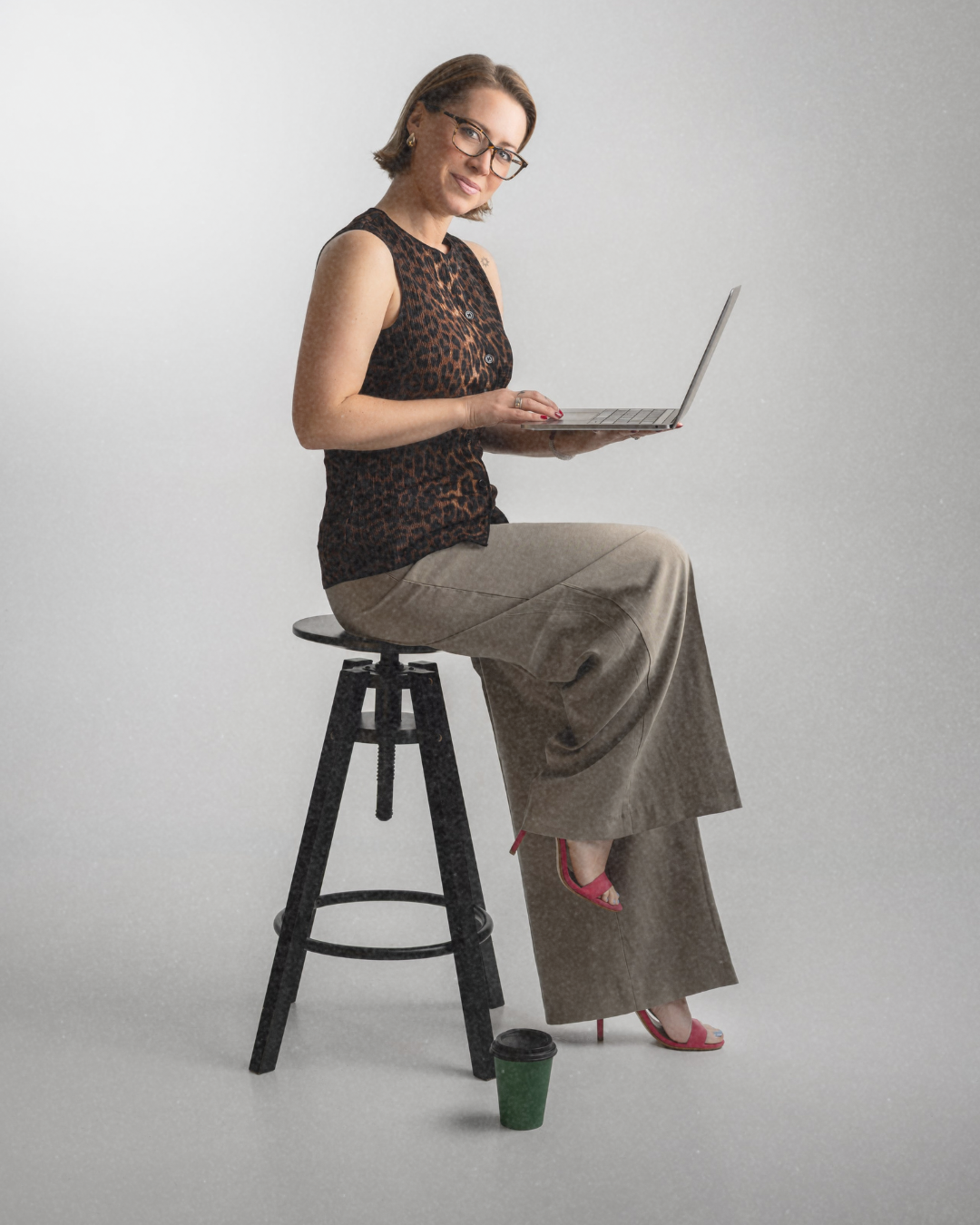 A woman sitting on a stool, holding a laptop, dressed in a sleeveless leopard print top and beige wide-leg pants, wearing glasses and red high-heeled sandals, with a green coffee cup on the floor.
