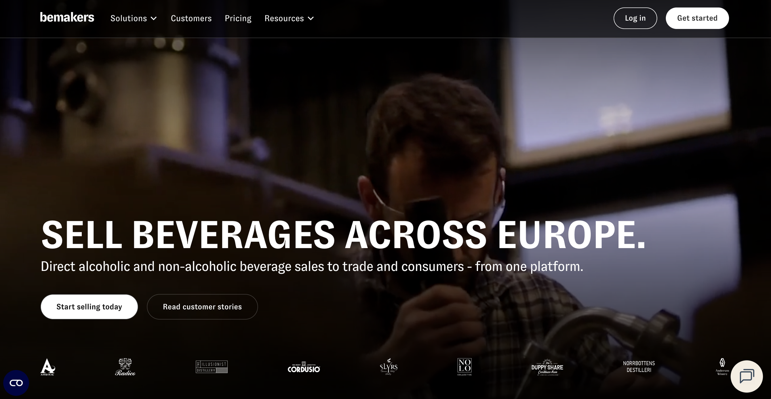 A person wearing glasses and a face mask pours a beverage from a tap at a bar or brewery, with a dimly lit background and multiple screens or monitors