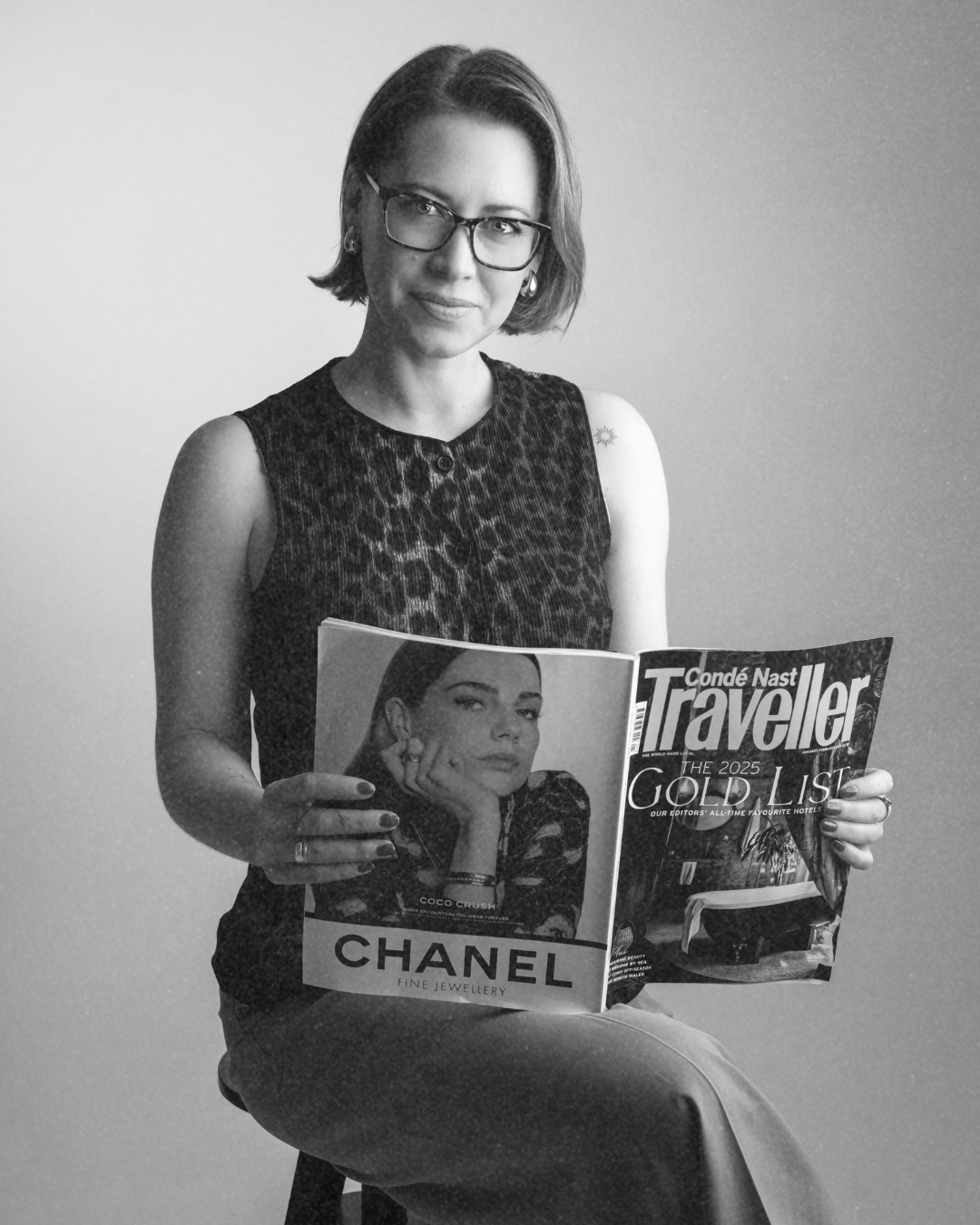 A woman with glasses and short hair sitting on a chair, holding an issue of Conde Nast Traveller magazine, wearing a sleeveless animal print top.