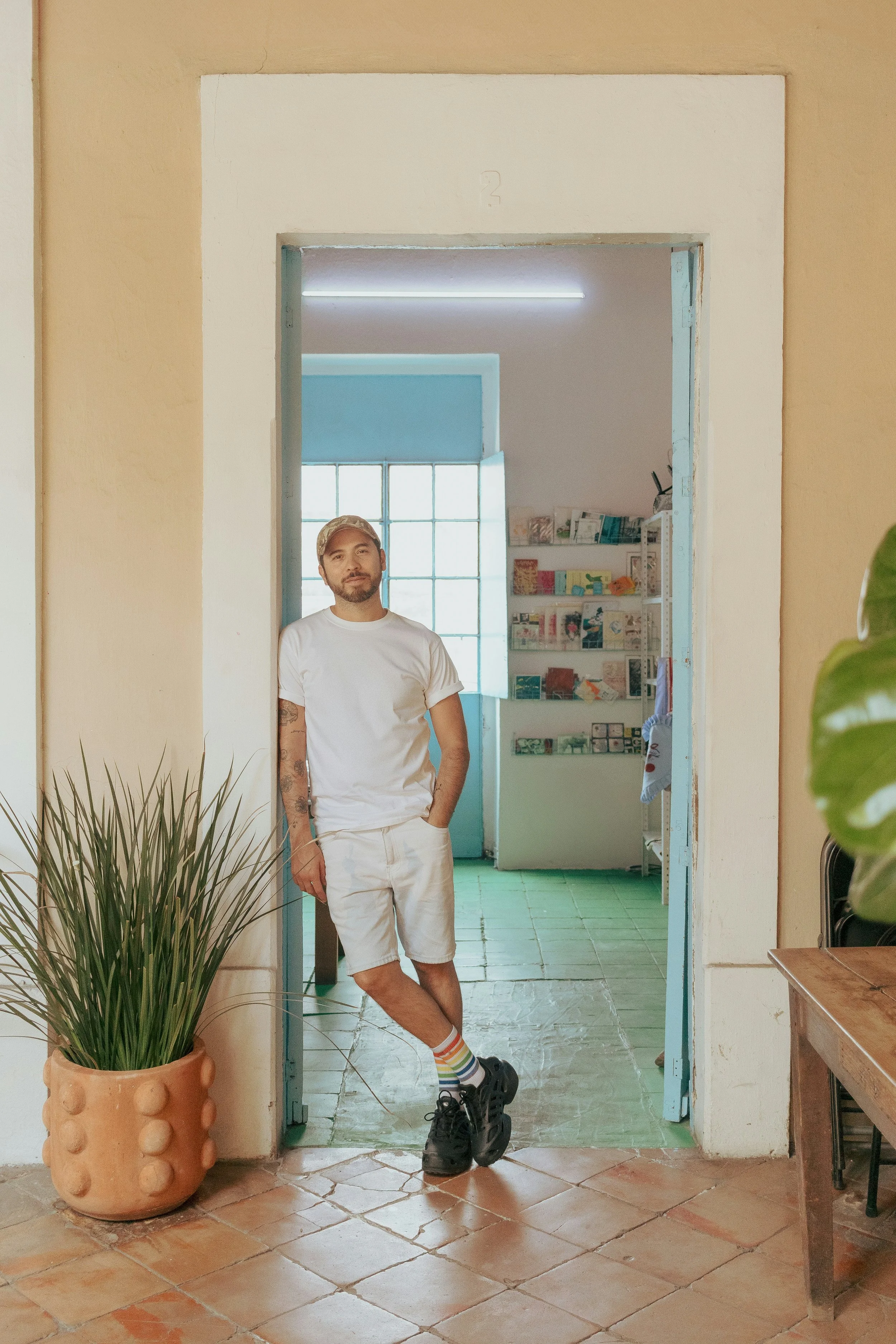 A man with a beard and tattoos wearing a white t-shirt, shorts, and rainbow socks, standing with one foot crossed over the other and leaning against a door frame inside a room with potted plants and a shelf of books.