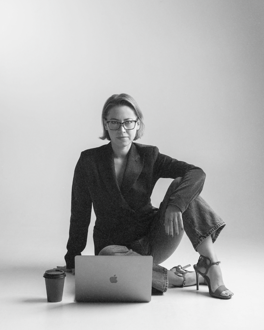 Black and white photo of a woman sitting on the floor with a laptop, a coffee cup, wearing glasses, a blazer, jeans, and high heels, against a plain background.