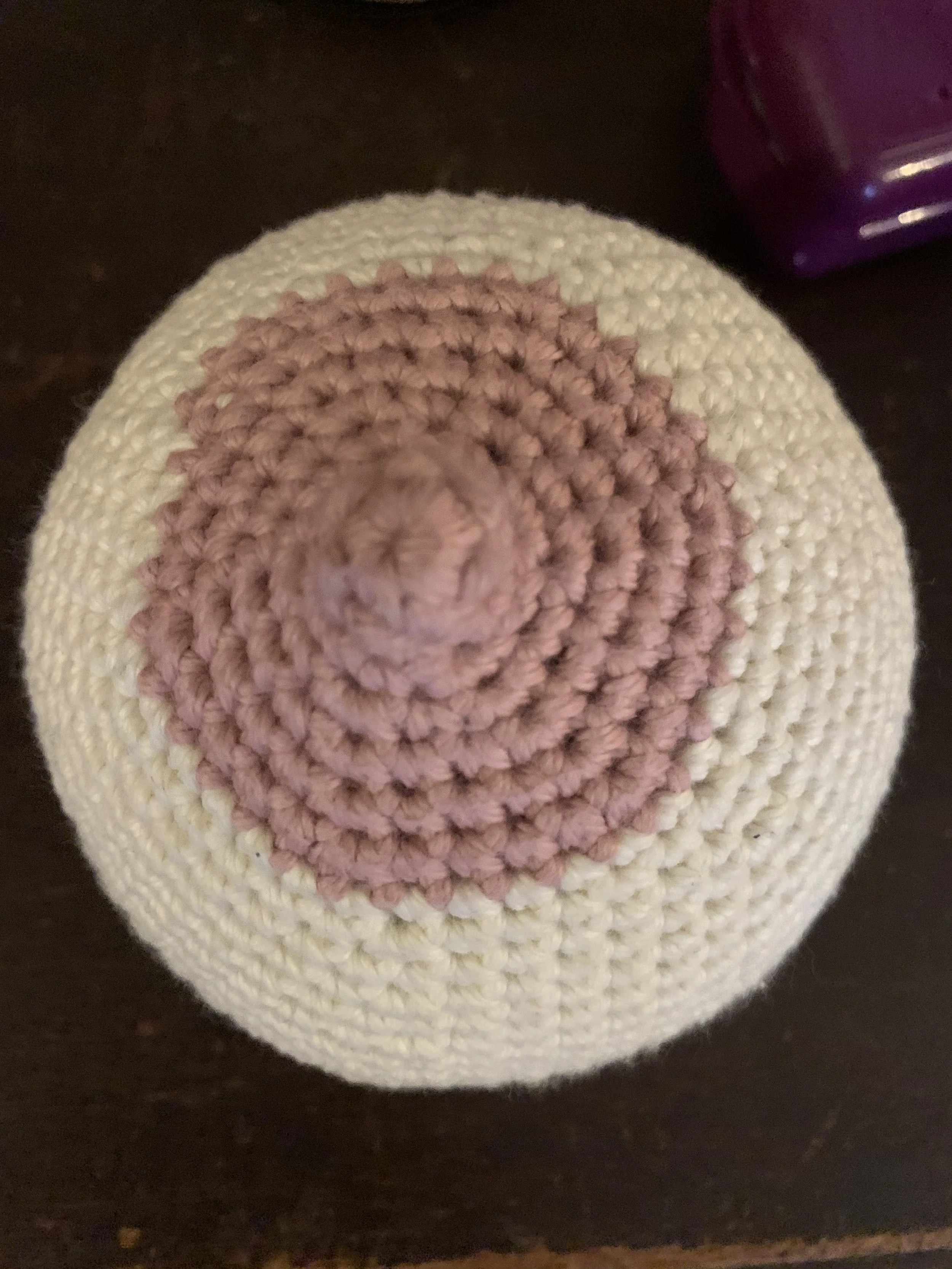 Knitted spherical object with concentric circles in beige, light brown, and pink colors, placed on a dark surface with a purple item partially visible in the background.
