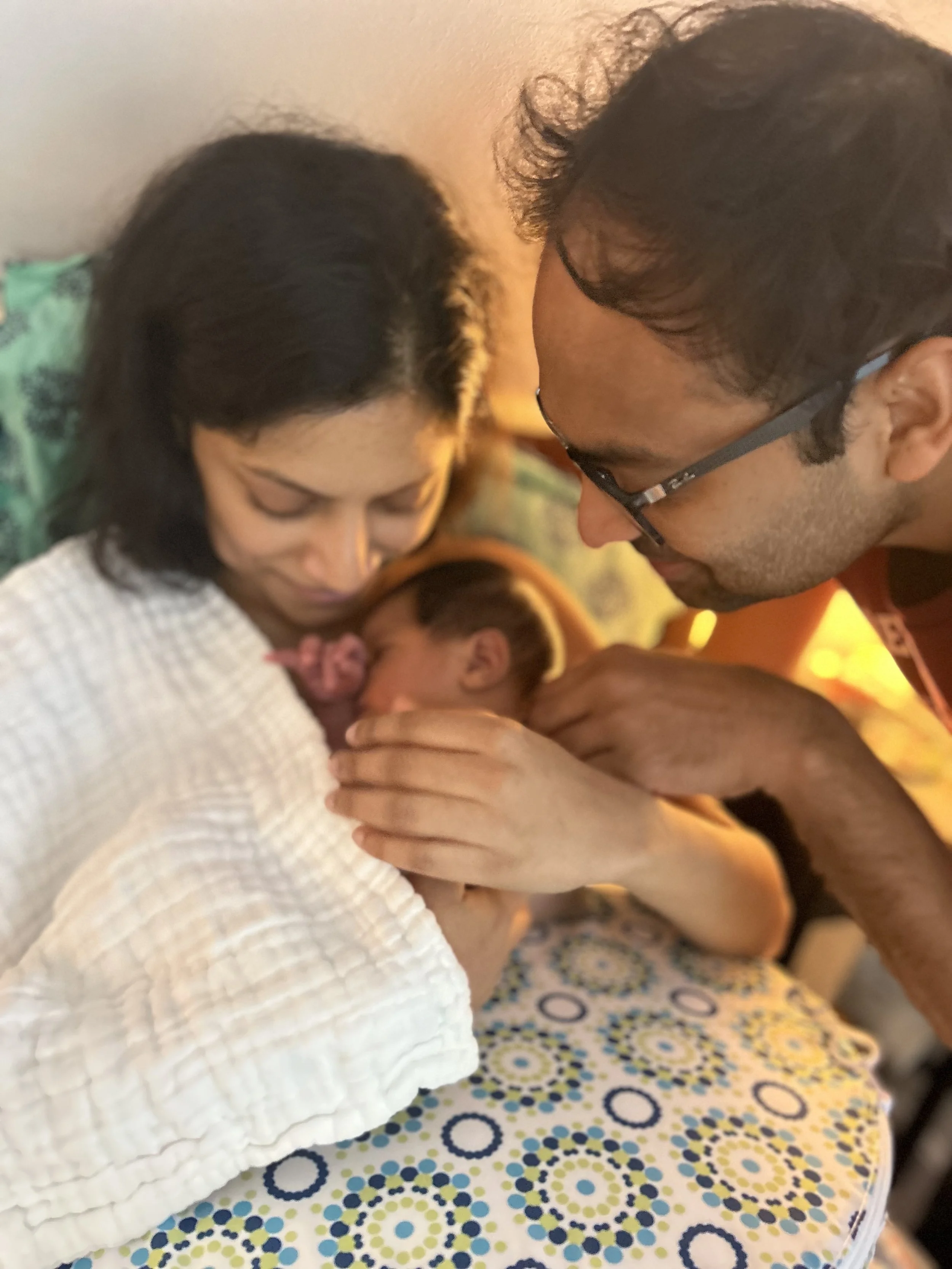 A woman holding a newborn baby close to her chest, a man with glasses gently touching the baby, in a warm, intimate moment.