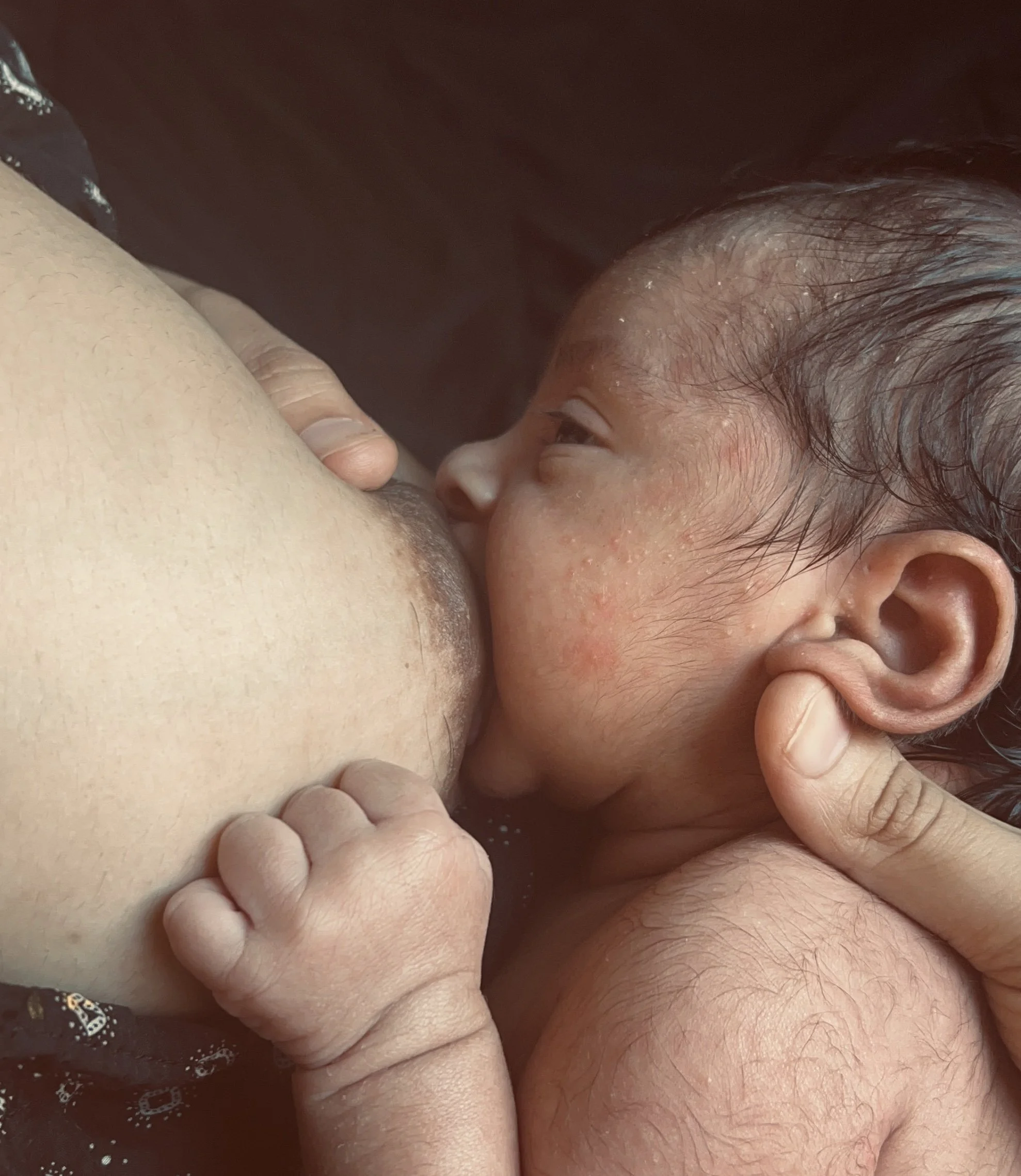 A baby is breastfeeding from a woman, with the baby's hand resting on the woman's breast and the woman gently holding the baby's head.