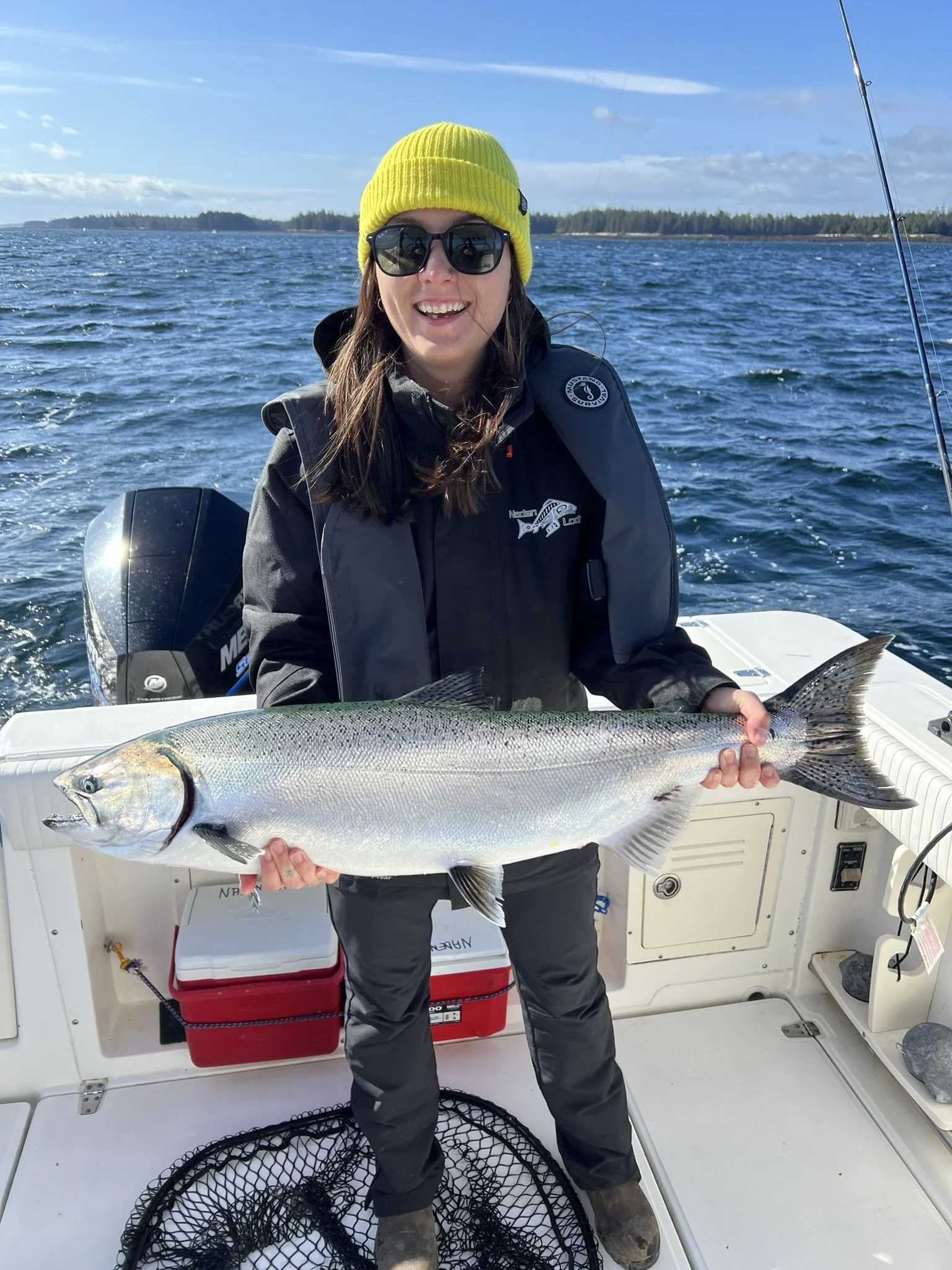 A woman in outdoor clothing, wearing a yellow beanie and sunglasses, smiling and holding a large fish on a boat in open water with land and blue sky in the background.