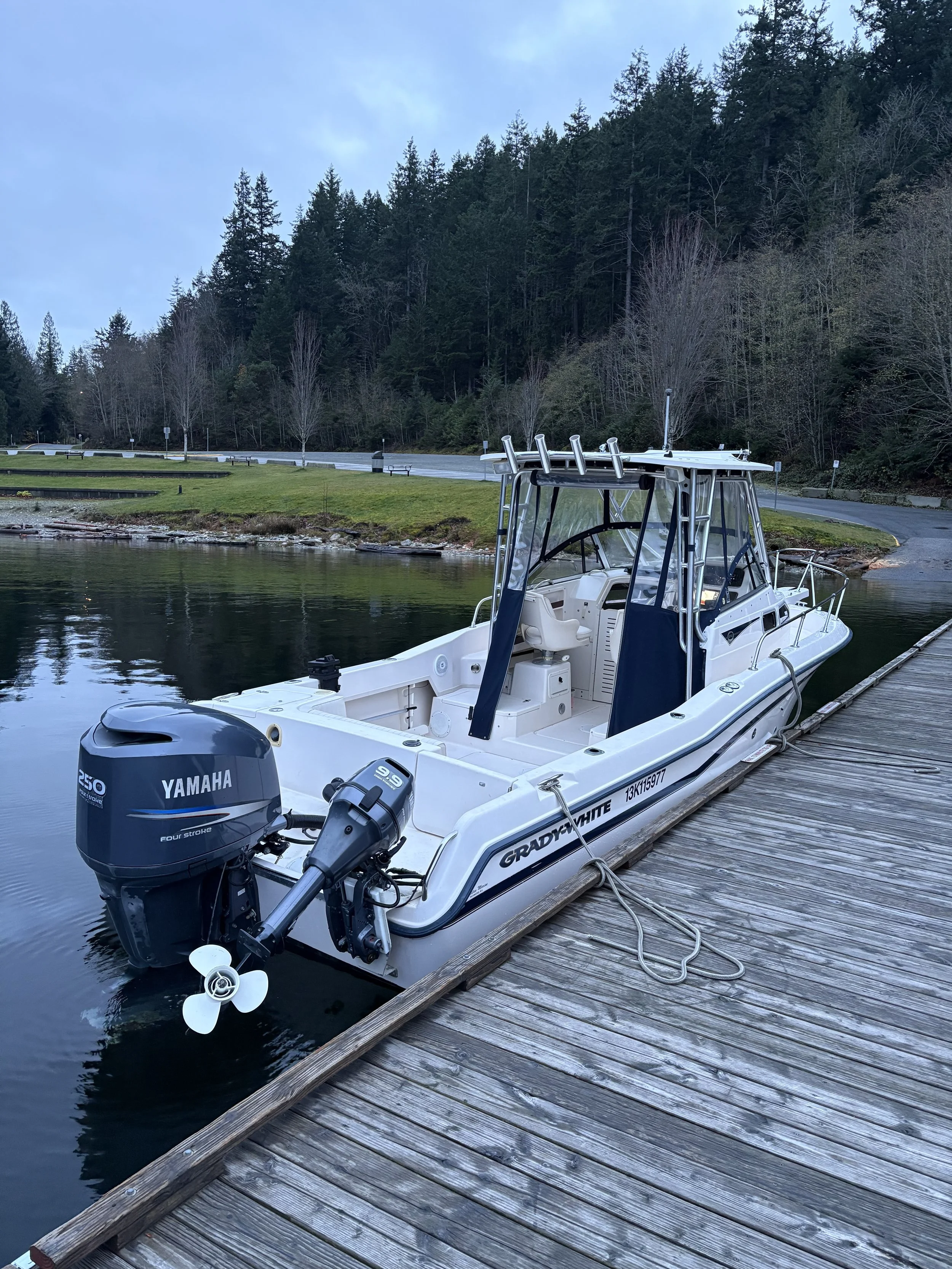 A white boat docked at a wooden pier on a calm body of water with a forested hillside in the background.