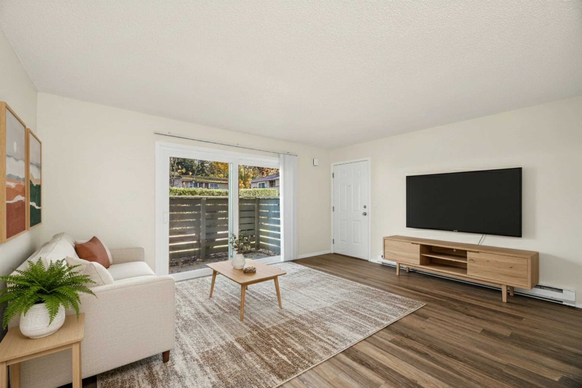 Living room with beige sofa, wooden coffee table, decorative artwork on the wall, sliding glass door with outdoor view, wooden TV stand, flat-screen TV, plants, and hardwood floors.