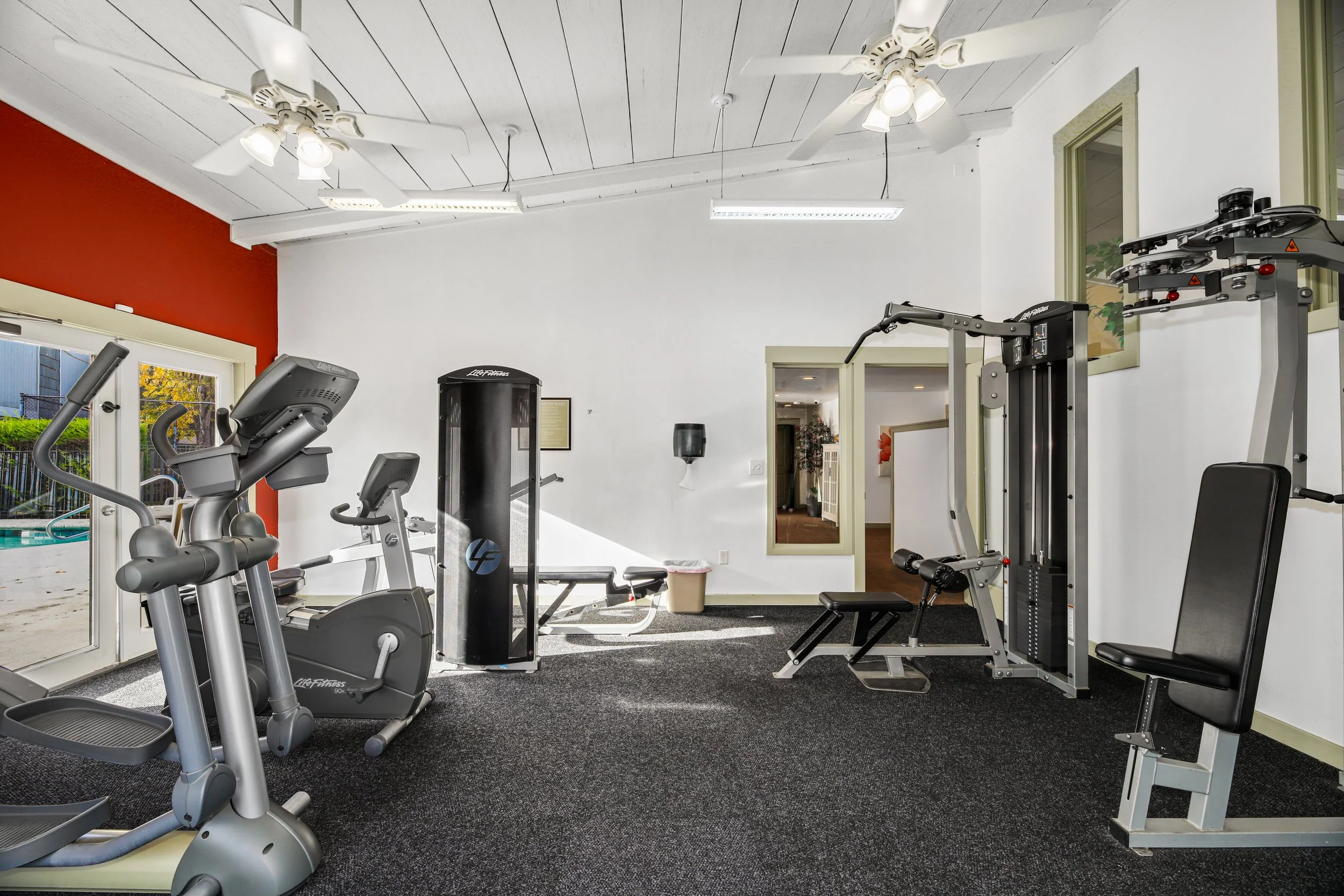 Empty gym room with exercise equipment including a treadmill, elliptical, weight machine, and bench, with a door to an outdoor pool area on the left side and a window on the right.