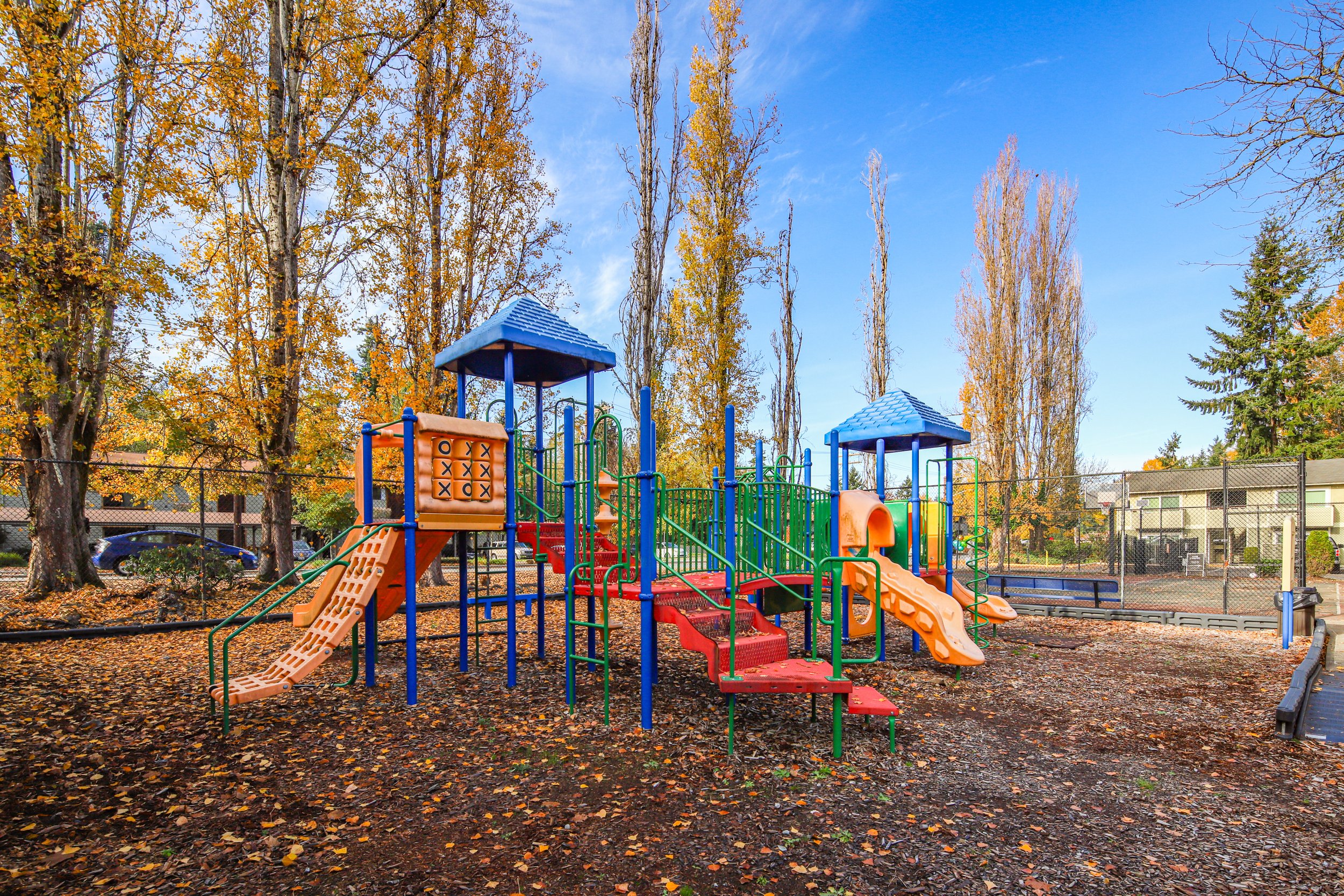 Colorful playground equipment with slides and climbing structures in a park during autumn, surrounded by trees with yellow and orange leaves.