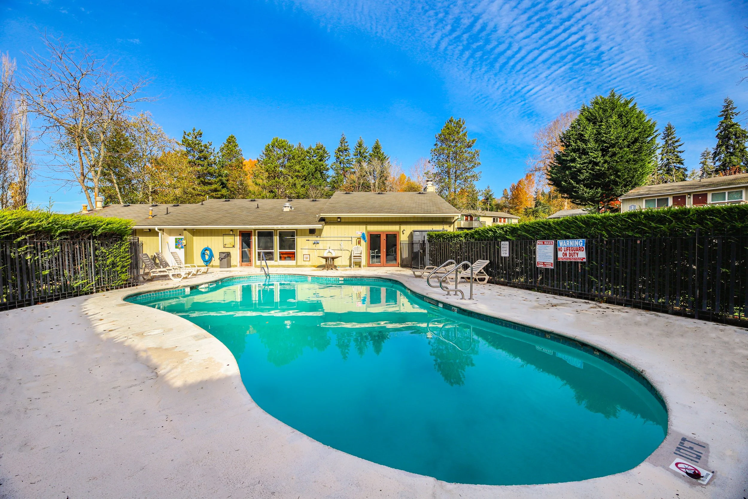 A backyard swimming pool with a concrete deck, lounge chairs, and a safety fence, with a house and trees in the background under a partly cloudy blue sky.