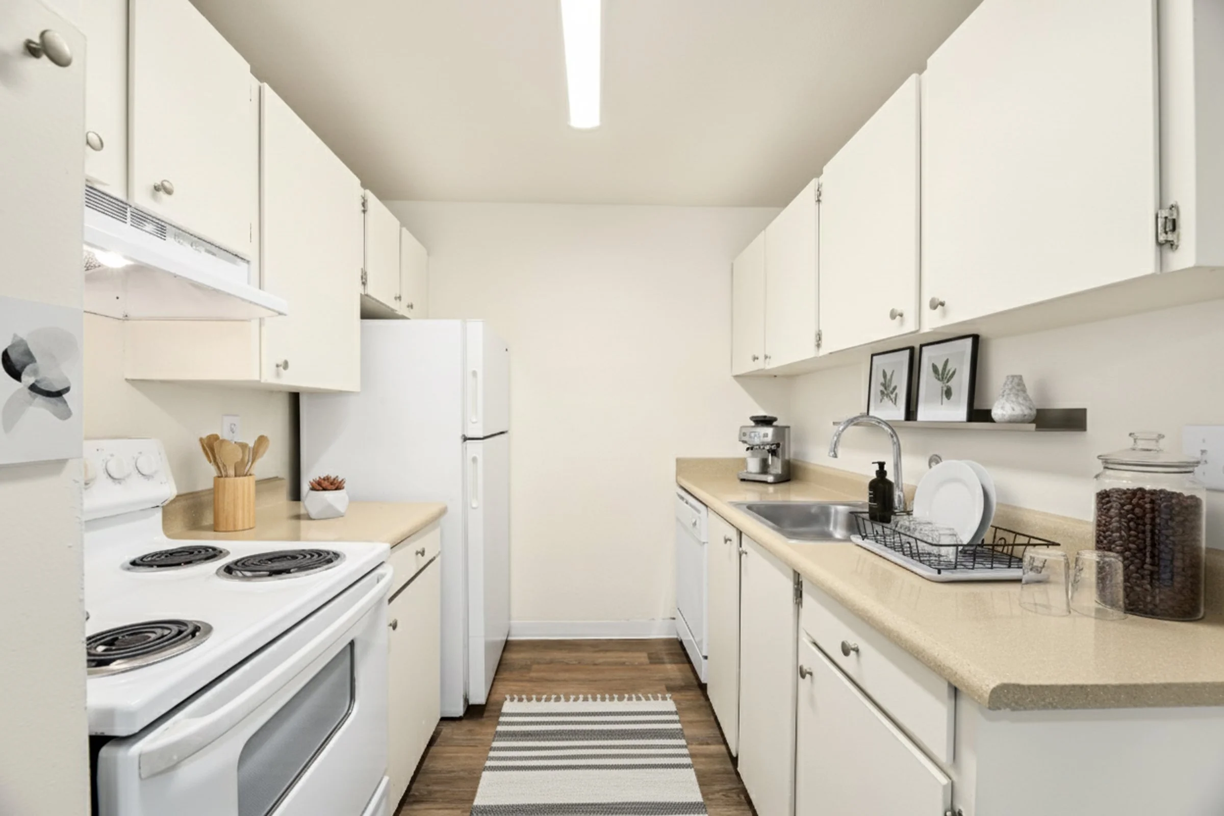 A compact kitchen with white cabinets, a white stove, a white refrigerator, and beige countertops. The kitchen has a small rug on the wood floor, a coffee maker, a dish drying rack with dishes, and decorative items such as framed botanical prints and a vase.