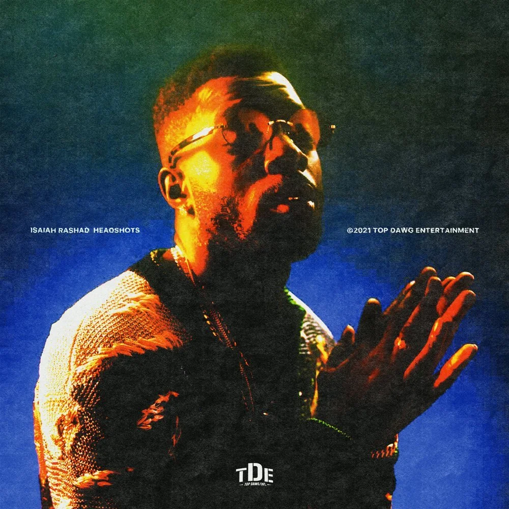 A portrait of Isaiah Rashad wearing sunglasses with hands clasped near his face, illuminated with colorful lighting, against a gradient background.