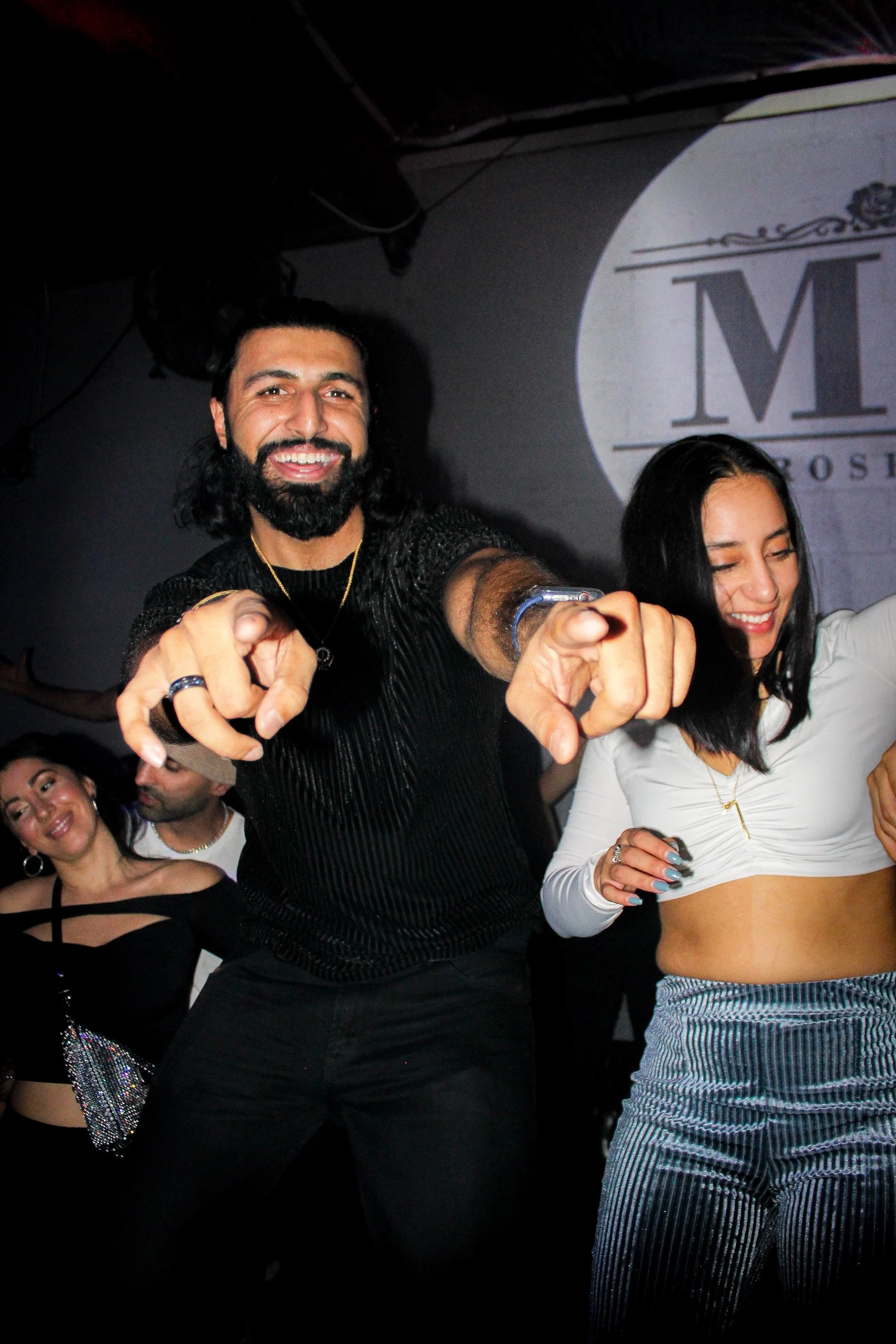 People dancing and smiling at a party, with a man pointing towards the camera and a woman next to him, in a lively social setting.