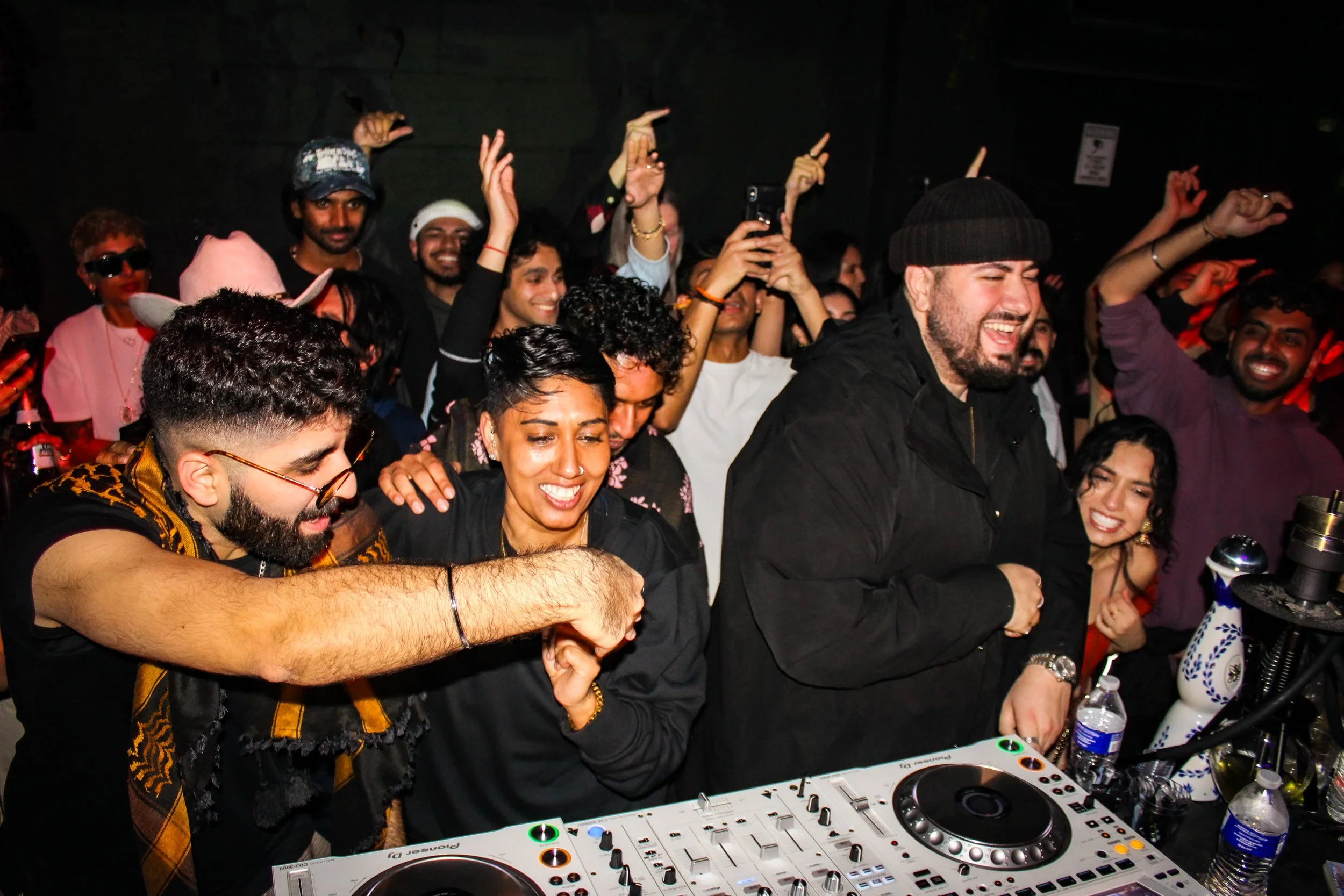 A lively group of people dancing and celebrating at a party with a DJ booth in the foreground, smiling and enjoying the moment.