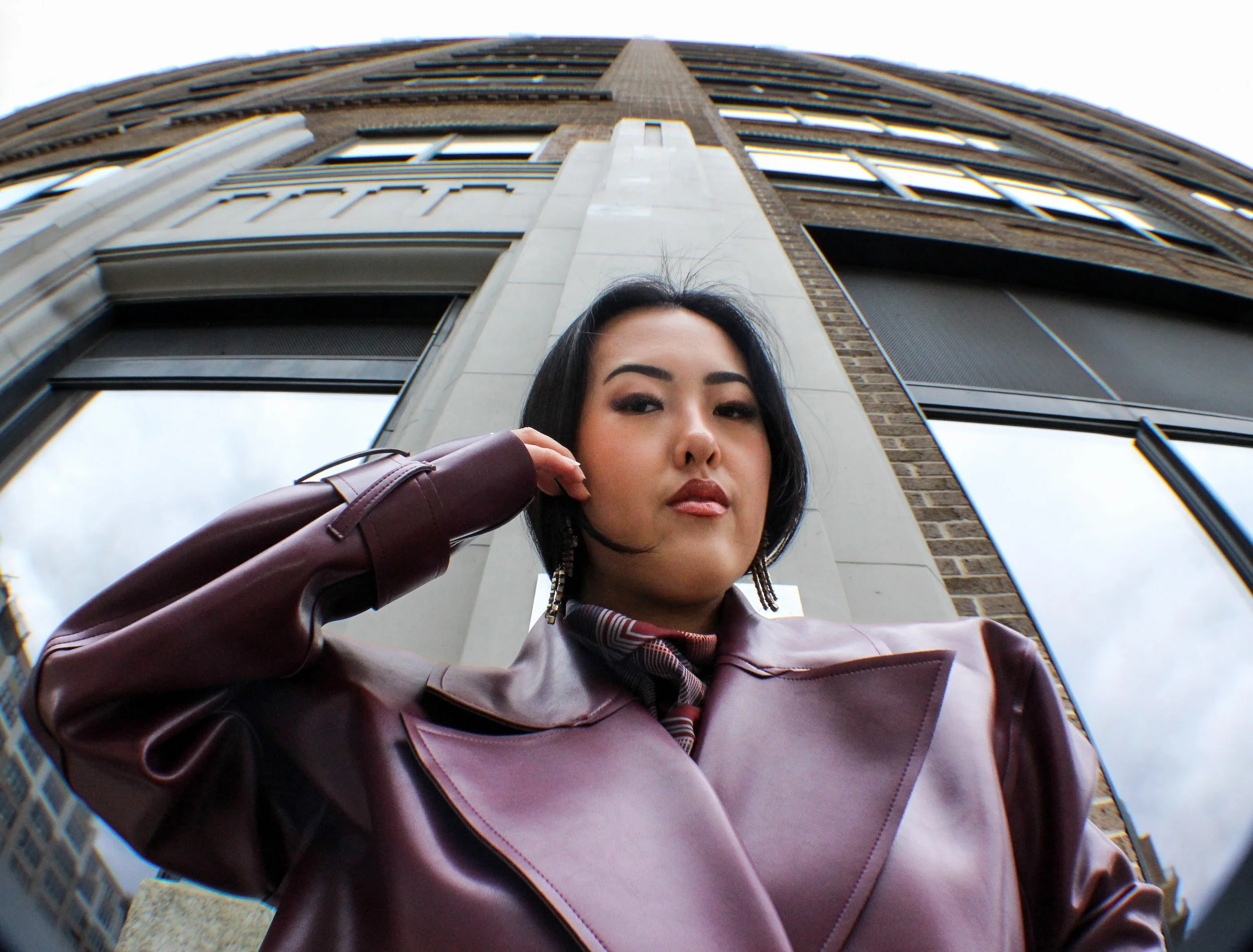 A woman with black hair and wearing a burgundy leather jacket poses outdoors in front of a tall building, looking into the camera, using a fisheye lens that makes the building appear curved.