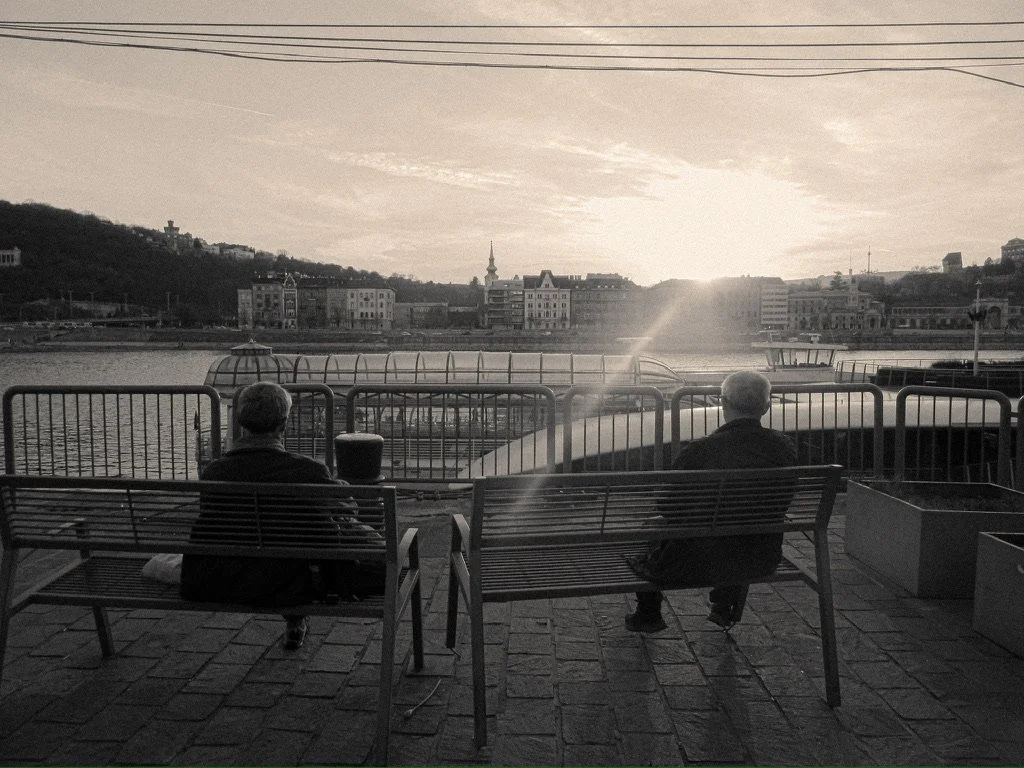 Two men sit on benches facing a river, with a railing in front of them, during sunset in a cityscape.