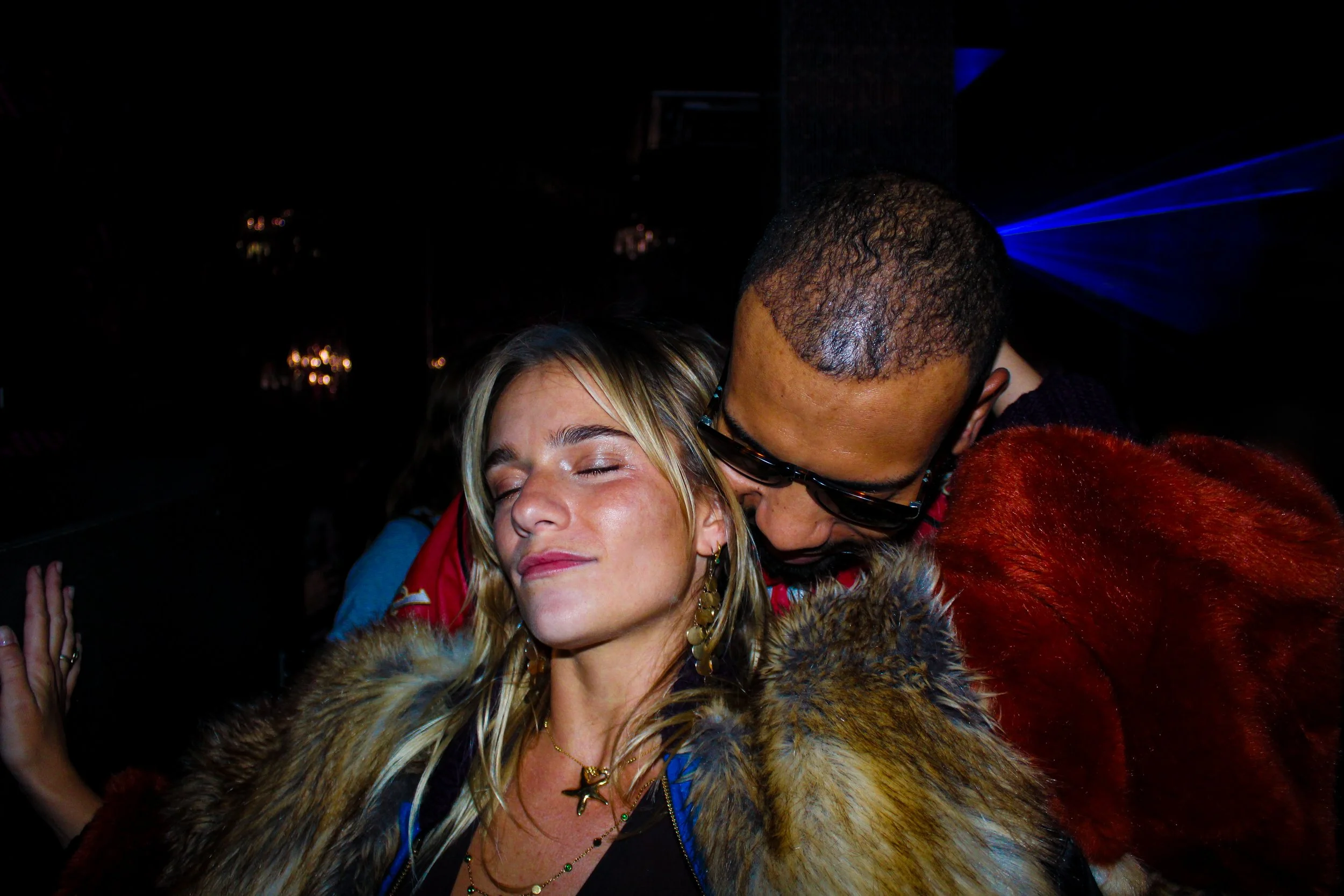 A woman with blonde hair and closed eyes appears to be enjoying a moment as a man in sunglasses leans in close, possibly kissing or whispering to her, in a dark, possibly nightclub setting with blue light effects.