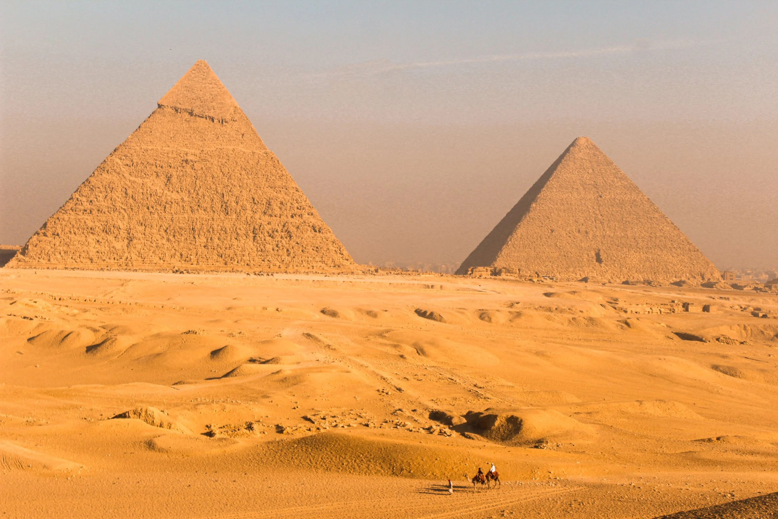 Two large pyramids in the desert with a person riding a camel in the foreground.