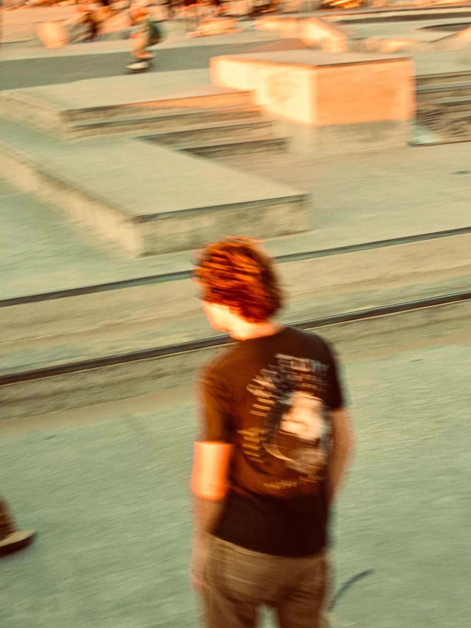 A person with curly hair wearing a black t-shirt and gray pants standing on a skatepark with skateboarding areas and ramps in the background.