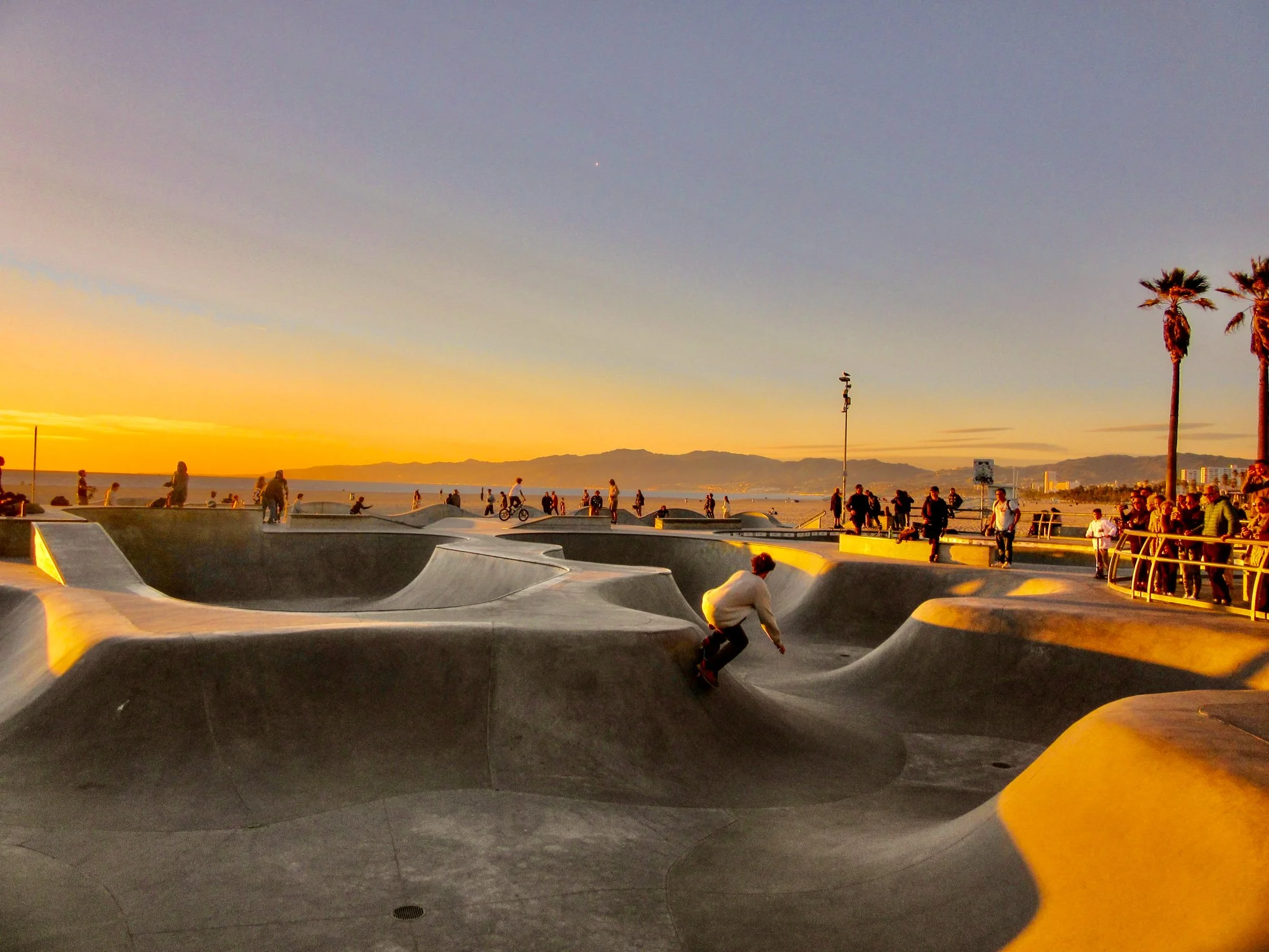 Skate park by the beach at sunset with many people skateboarding, biking, and walking, along with palm trees and mountains in the distance.