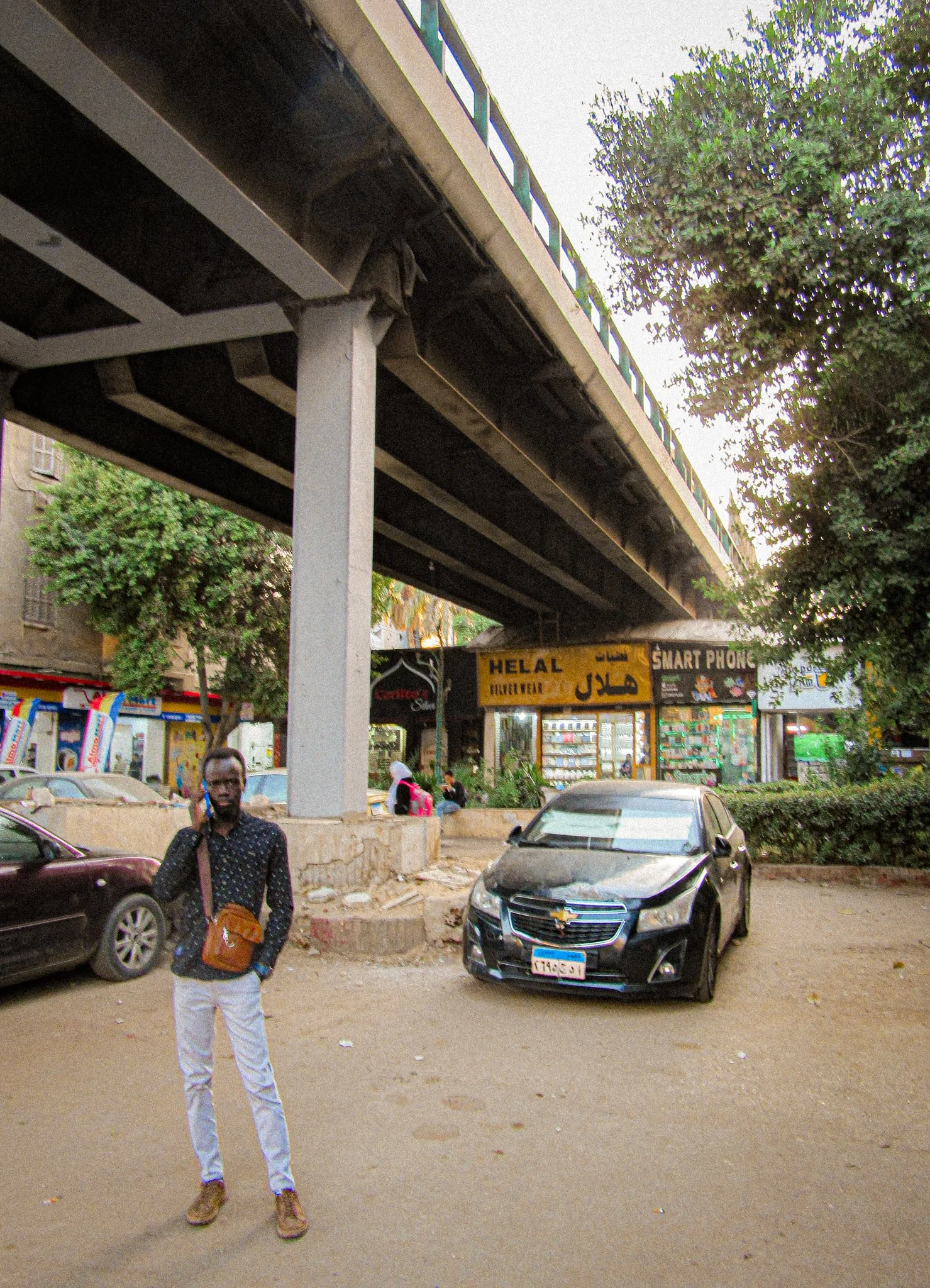 A man standing on a dirt parking lot under an elevated train track. There are parked cars, small shops with signs, trees, and a few people in the background.