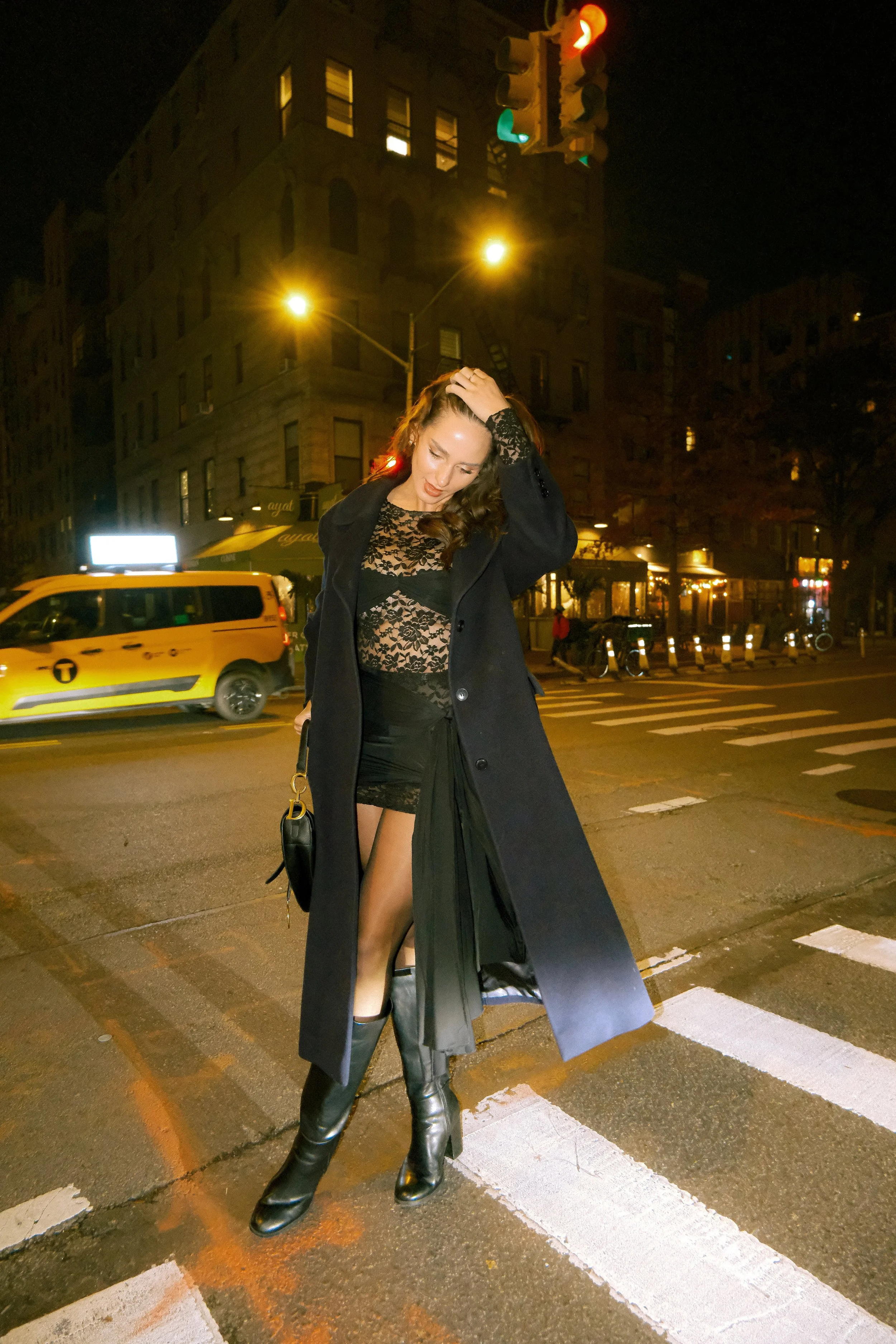 A woman with long hair standing on a city street at night, dressed in a black lace dress, black coat, and knee-high black boots, with her hand on her head and head slightly tilted.
