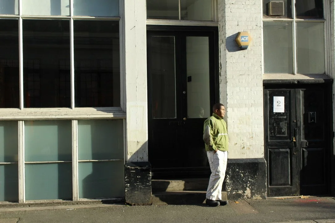 A man standing outside a building with large glass windows, a black door, and an ADF security alarm box on the wall, wearing a green jacket and white pants.
