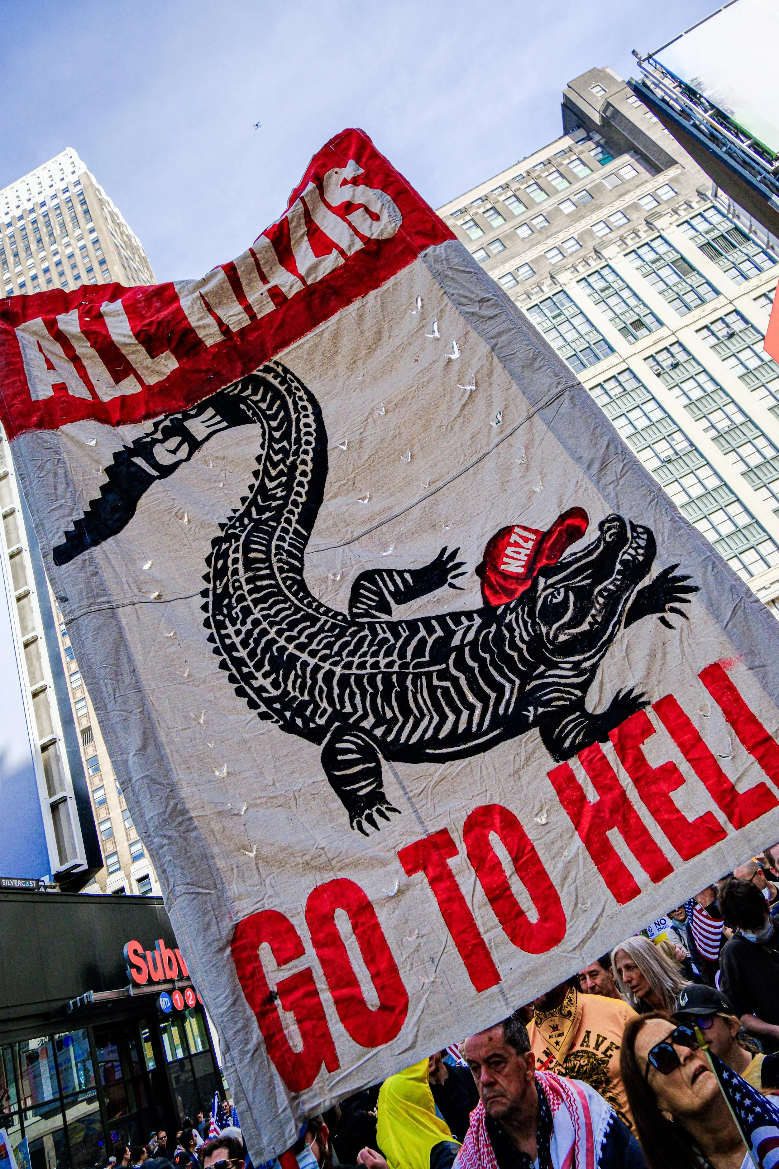 A protest or demonstration with a large banner depicting a crocodile wearing a red cap labeled "NAZI". The banner has the words "GO TO HELL" at the bottom and the partly visible word "ALLIES" at the top. The protest is in an urban area with tall buil