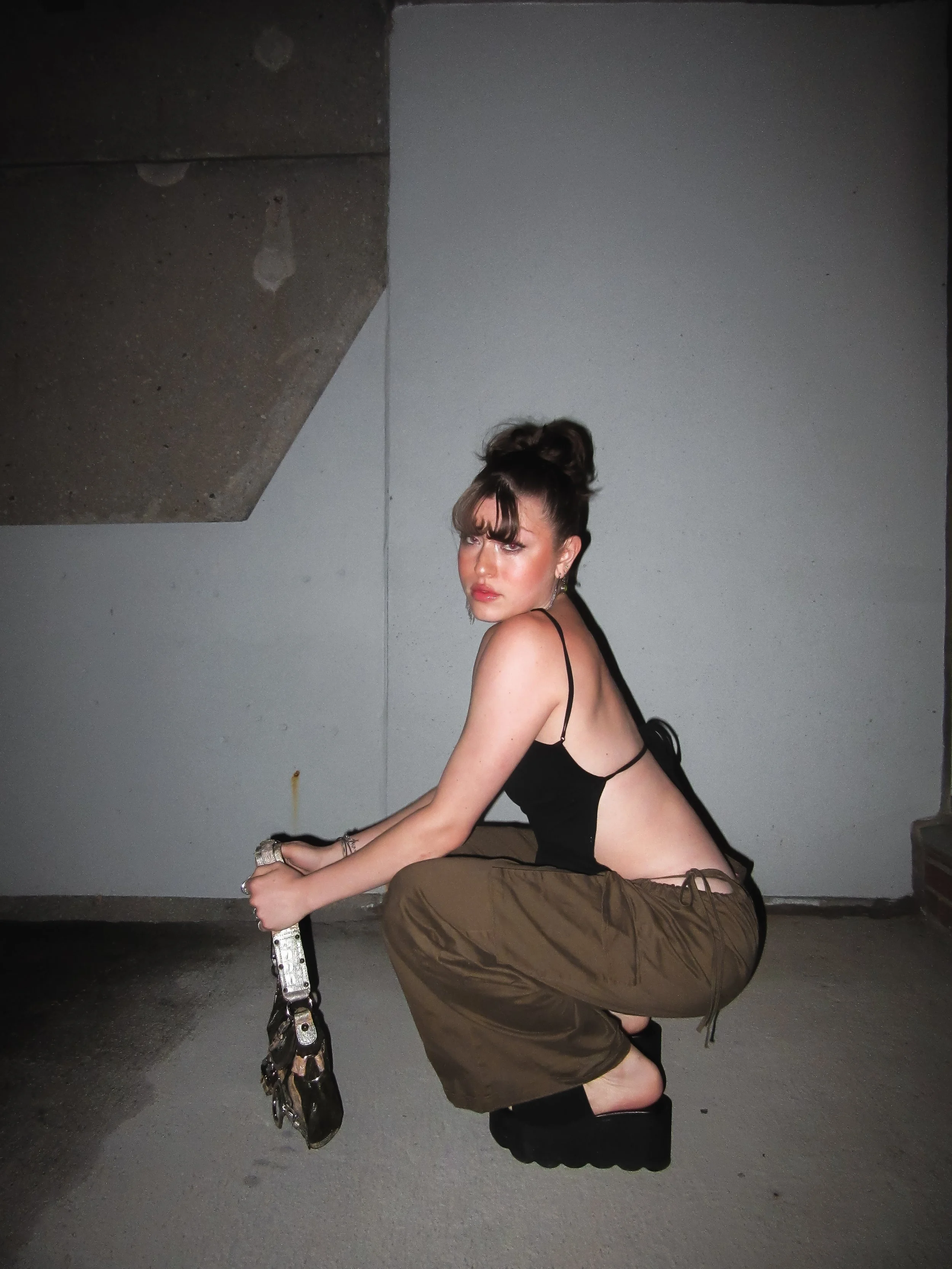 A woman with dark hair in a bun crouching on a concrete surface, wearing a black top with thin straps, loose brown pants, and black platform shoes, holding a purse, against a gray wall with a concrete ceiling in the background.