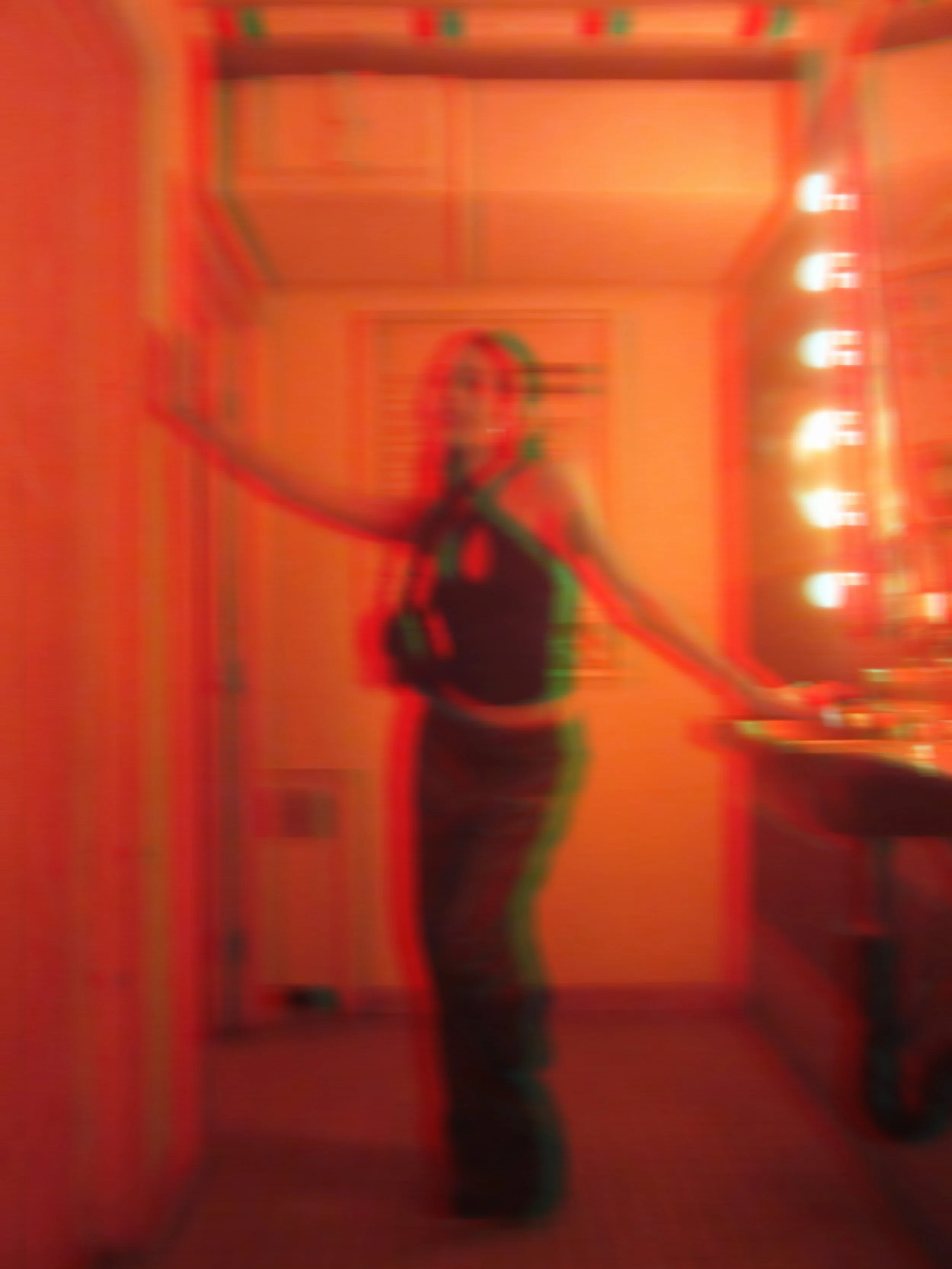Blurry photo of a woman standing in a colorful room with orange walls, neon lights, and a mirror.