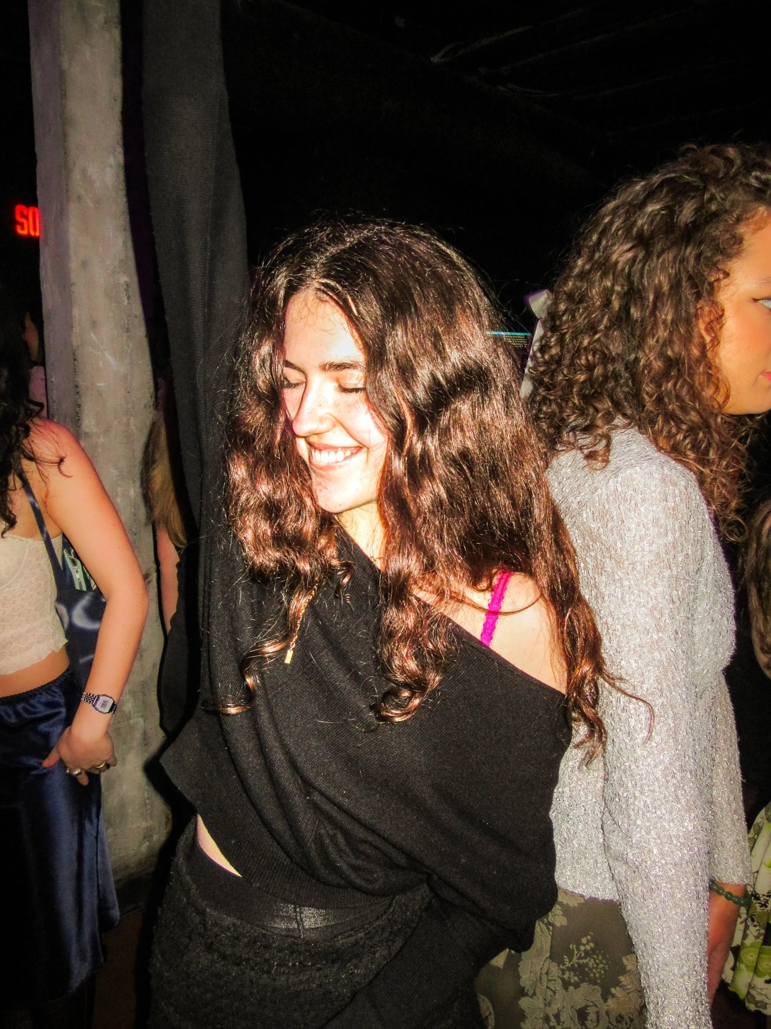 A young woman with long, curly brown hair smiling with eyes closed, wearing a black off-the-shoulder top and pink bra strap visible, standing at a party or social gathering.