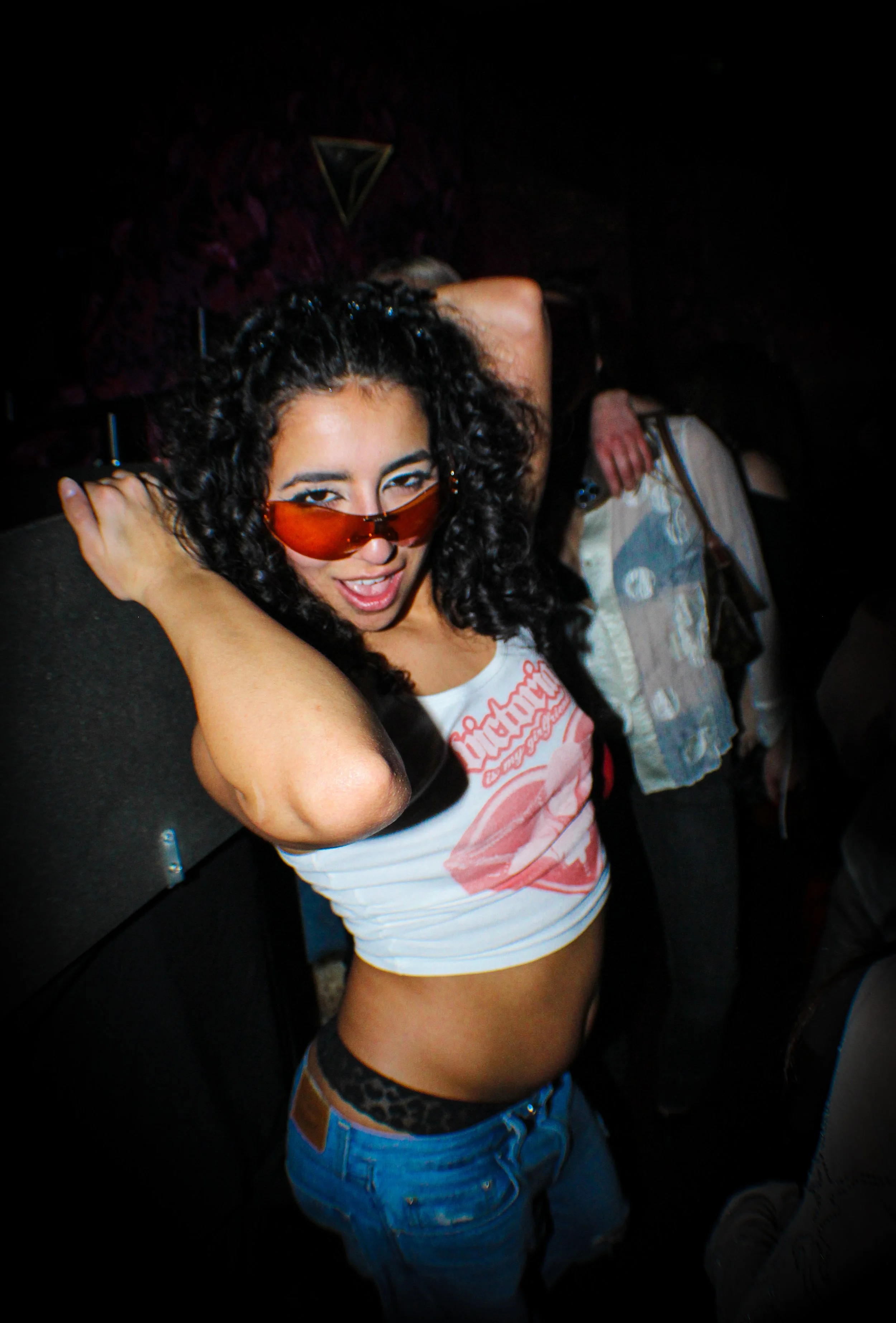 A young woman with curly hair wearing red sunglasses, a white graphic tank top, and blue jeans dancing at a party or nightclub.