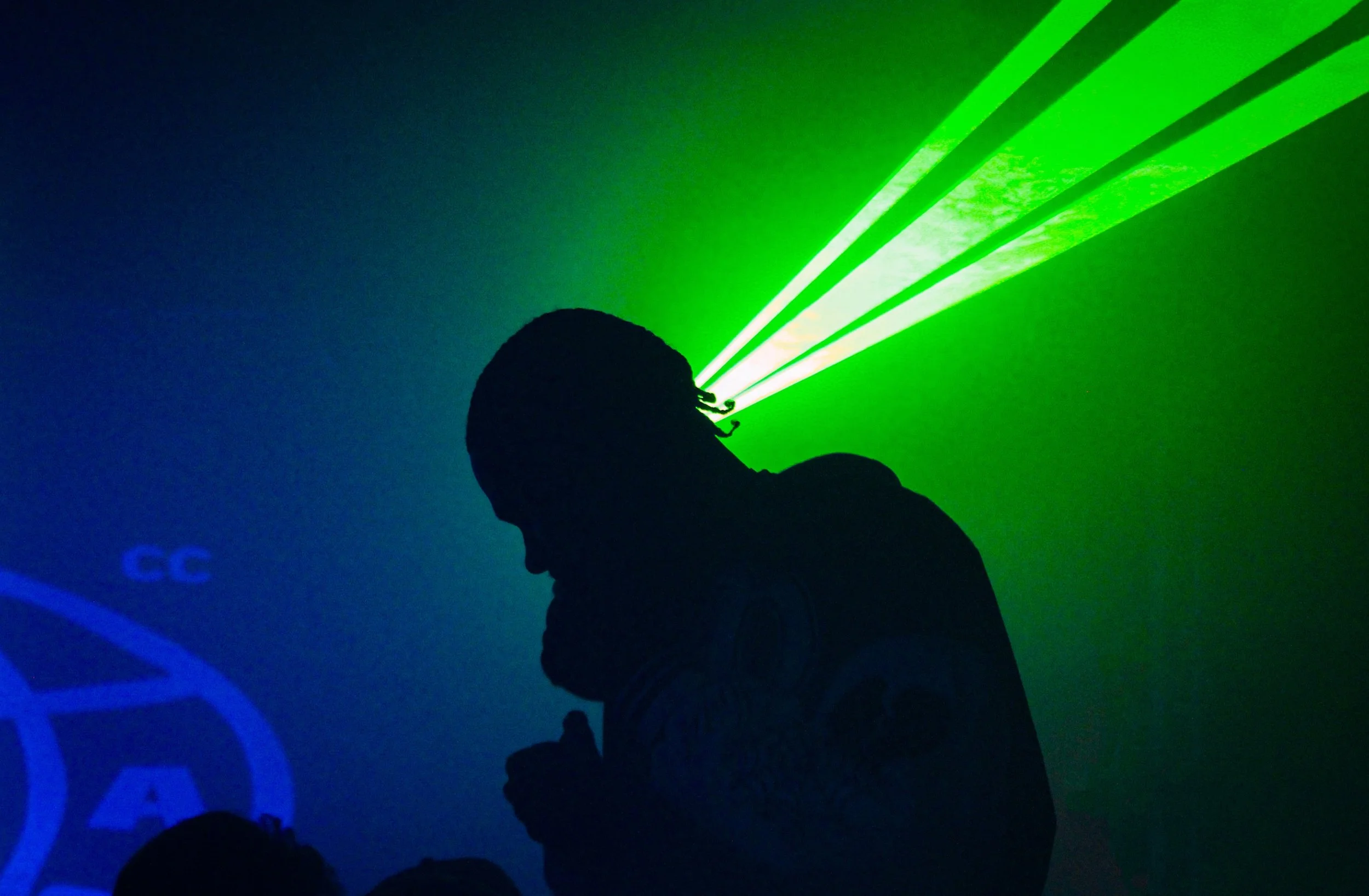 Silhouette of a person with dreadlocks in a dark room, illuminated by green and blue laser lights projection.