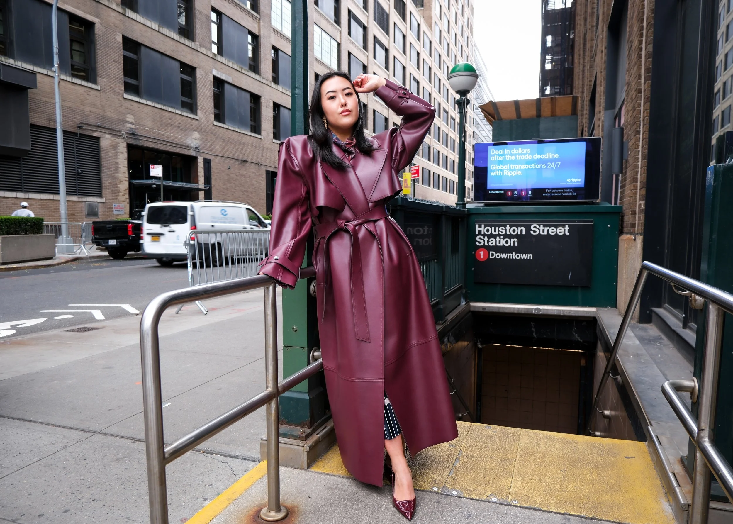 A woman in a long burgundy leather coat and heels standing at the entrance of Houston Street Station on a city street, with buildings, cars, and traffic visible in the background.