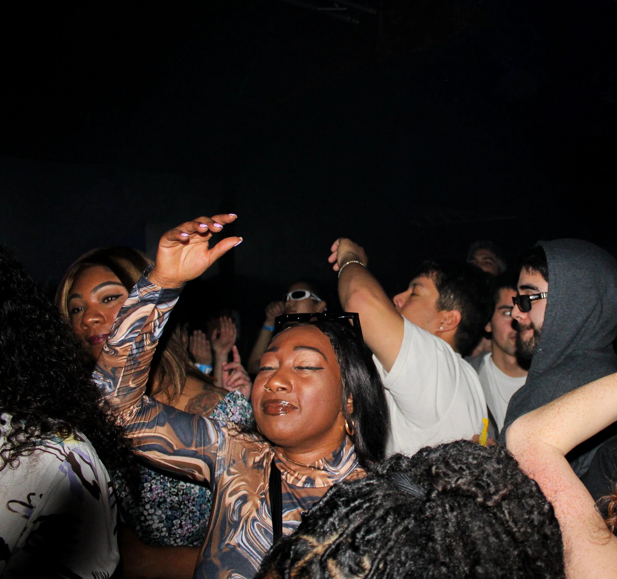 Crowd of diverse people dancing and enjoying music at a concert or party, with focus on a woman with closed eyes in the foreground raising her hand.
