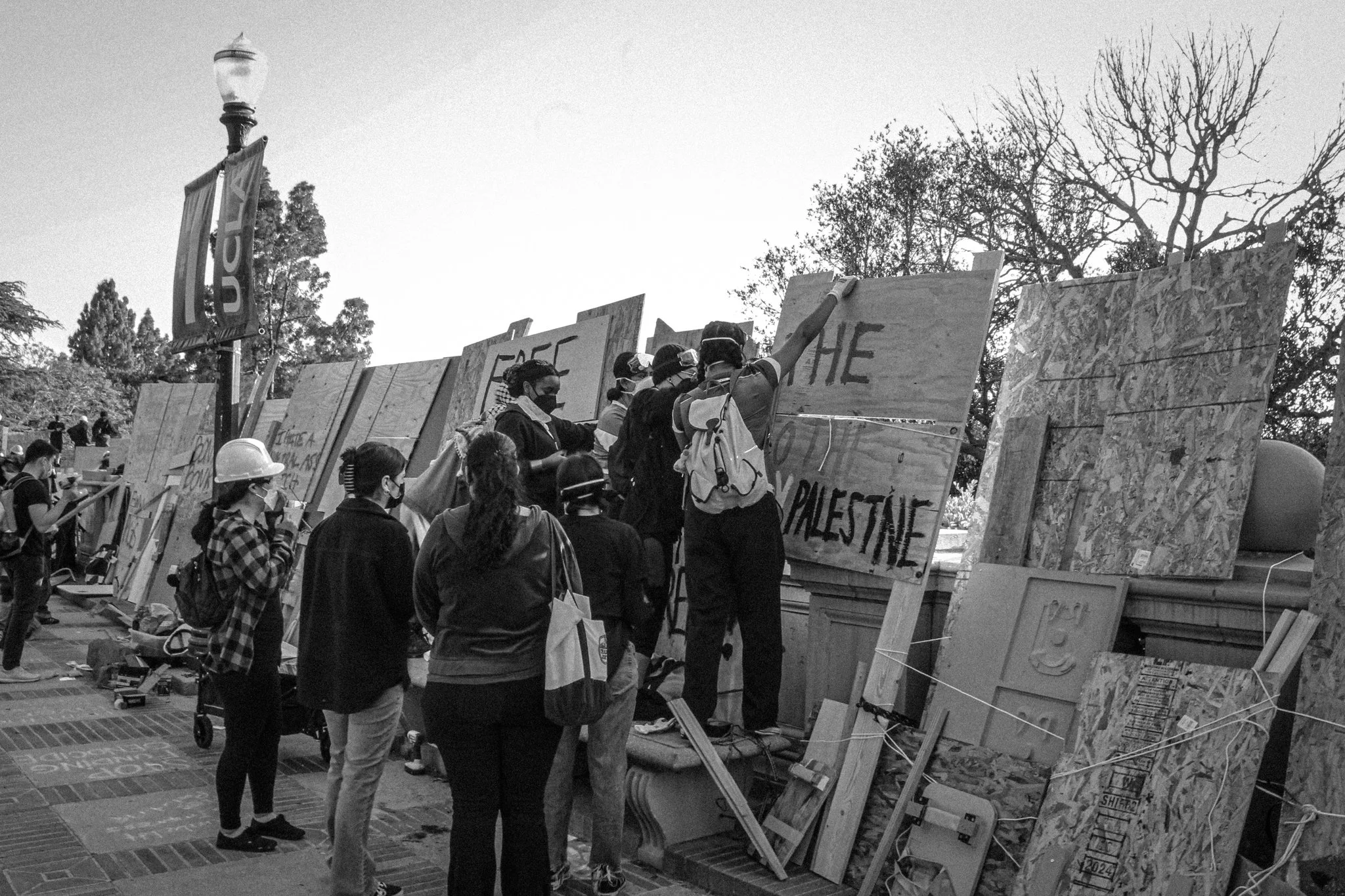 Group of protesters wearing masks and helmets, holding and standing near large wooden boards with messages, in an outdoor setting with trees.