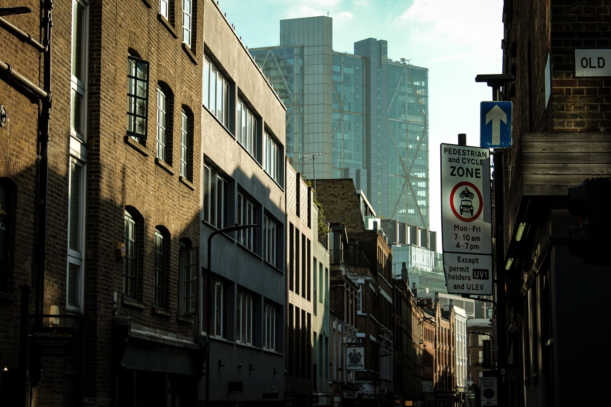 City street with tall buildings and a sign indicating pedestrian and cycle zone, with a directive for cars from Monday to Friday, 7-10 am, 4-7 pm.