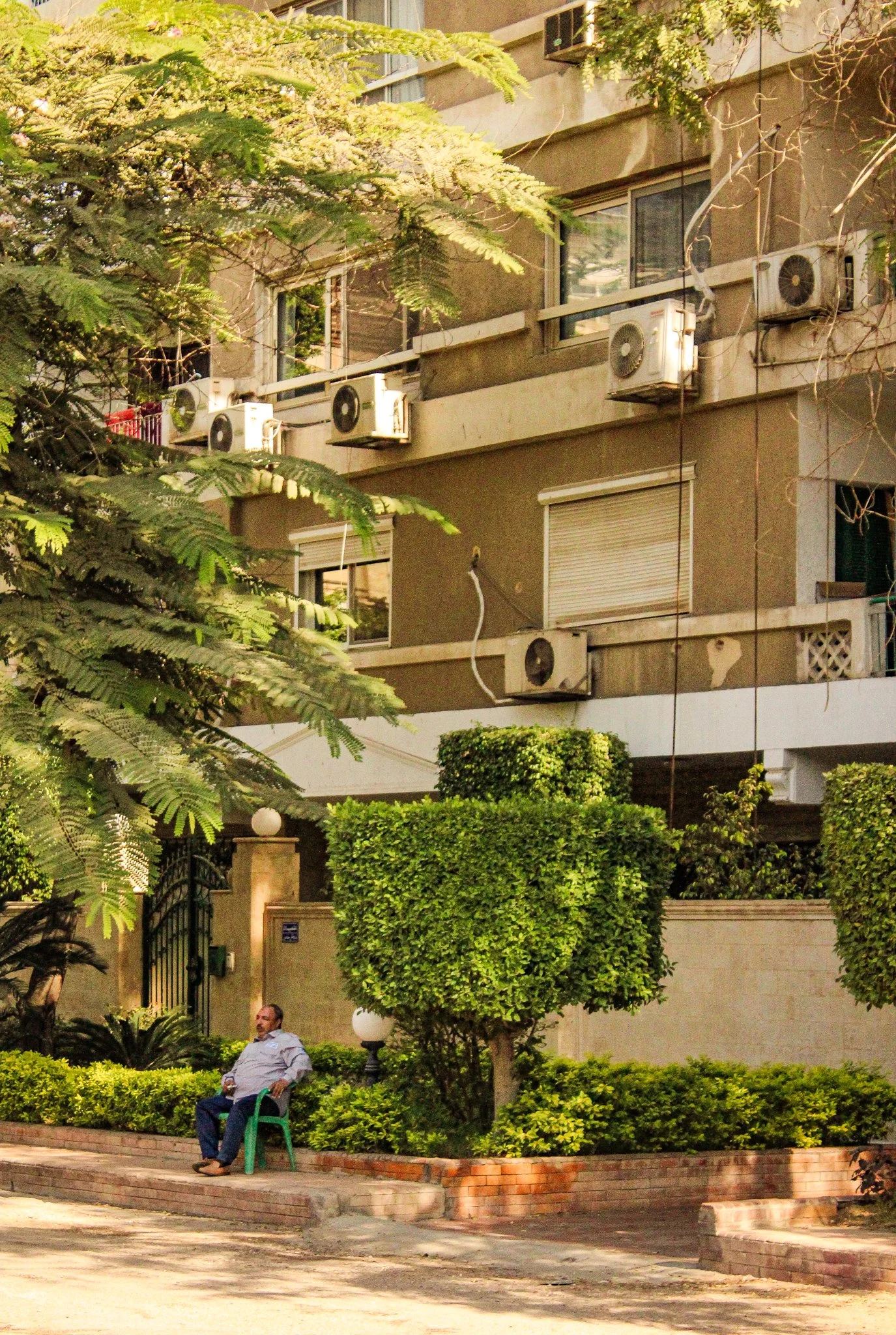 A man sitting on a green chair outside a gated building, surrounded by bushes and trees, with a multi-story residential building in the background that has several air conditioning units attached to the exterior walls.