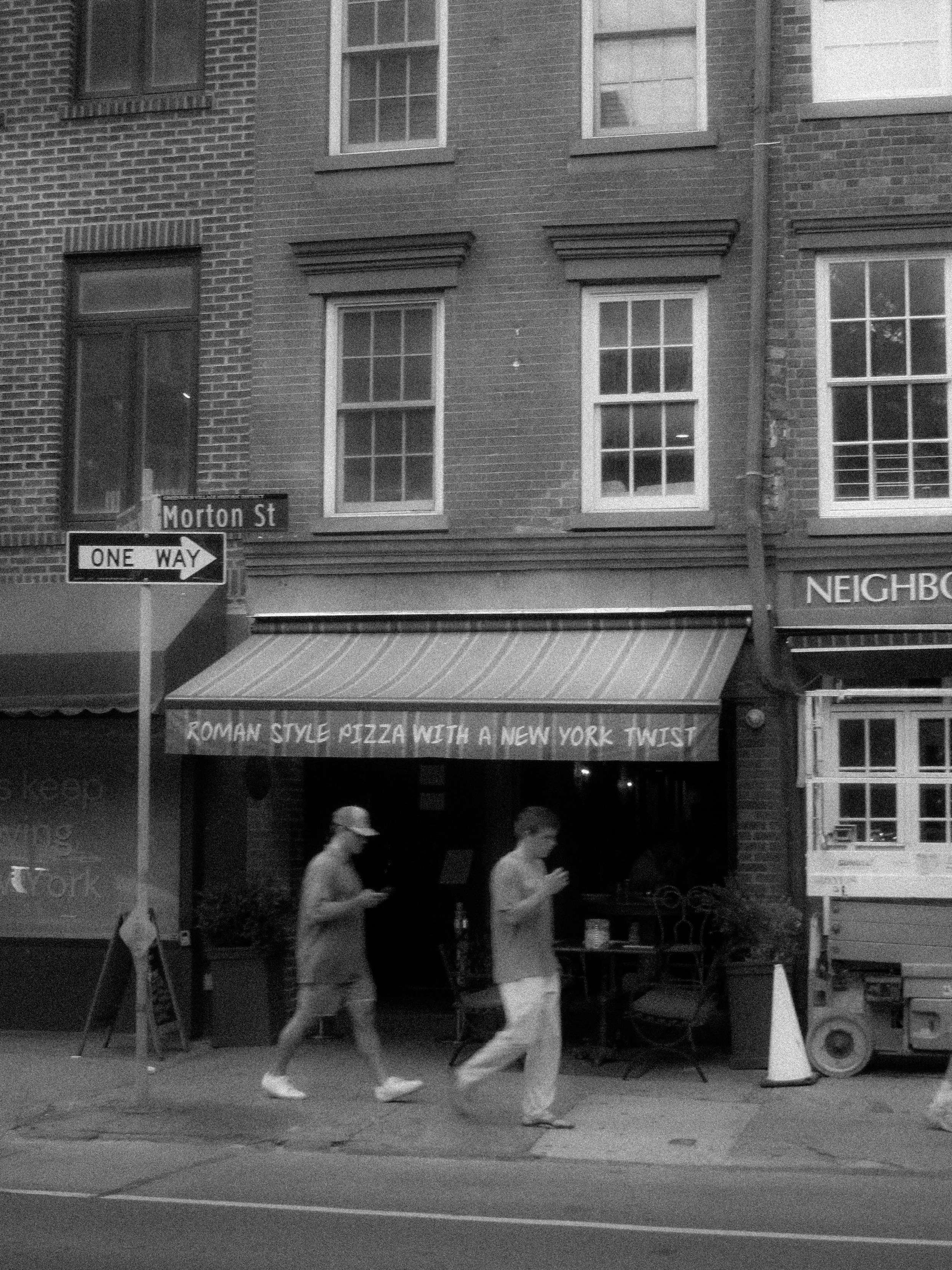 People walk past a pizza restaurant with a sign that reads 'Roman Style Pizza with a New York Twist' on Morton Street, in an urban area with brick buildings and a one-way street sign.